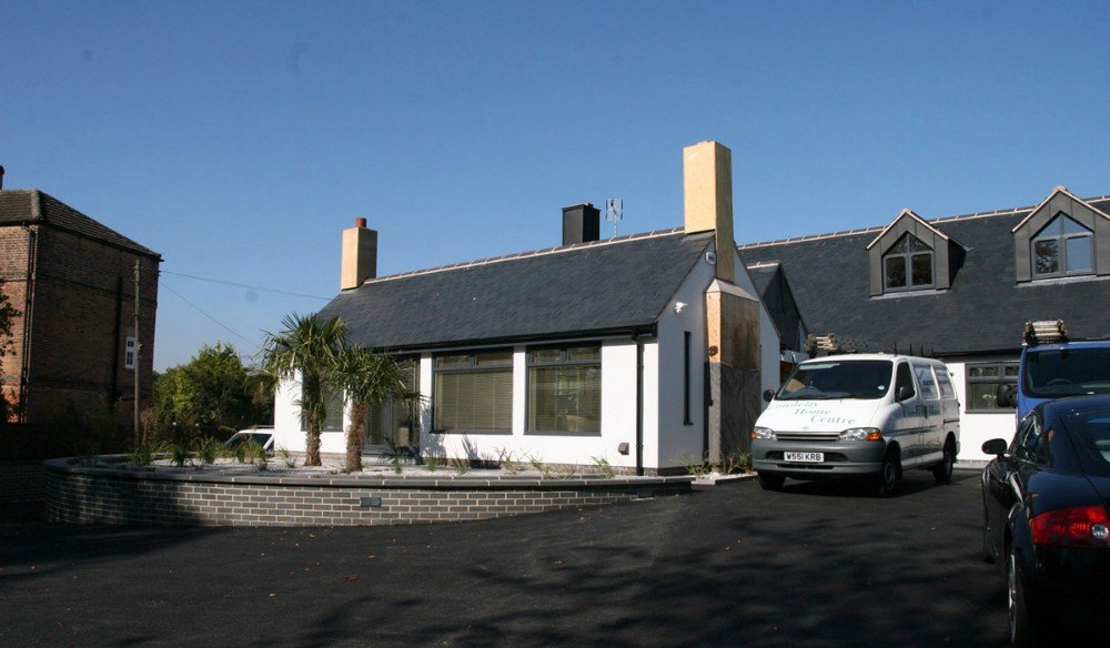 A small white house with a gray roof and tall chimneys, surrounded by a brick-bordered garden with palm trees, parked in front of a black asphalt lot, under a clear blue sky.