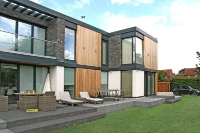 Modern two-story house with large windows, wooden and dark stone exterior, patio with lounge chairs, and a grassy yard.