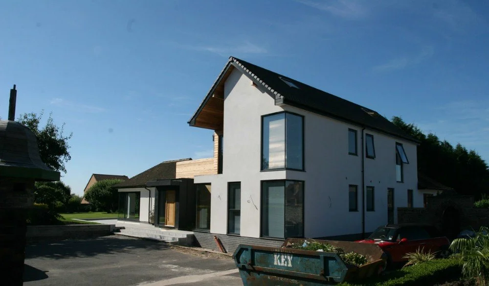 Modern two-story house with white exterior walls, large windows, and a black roof, located in a suburban neighborhood.