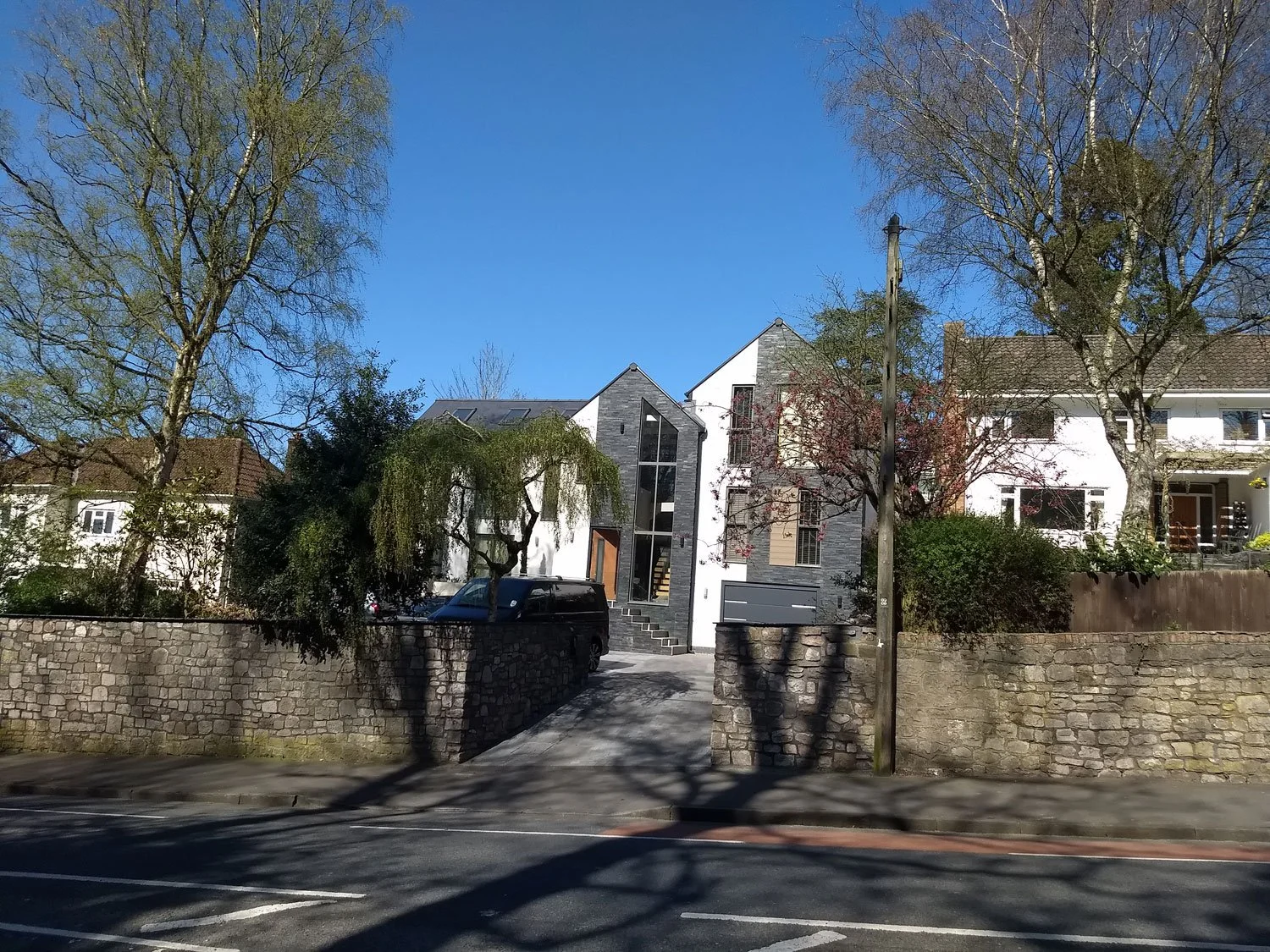 Modern multi-story house with black stone exterior, large glass windows, and a front staircase, surrounded by trees and a stone wall, with parked black van in driveway and a clear blue sky.