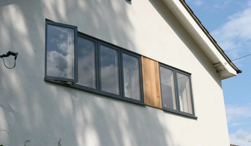Close-up of a side of a white house with a large window that has multiple panes, some of which are open, and a small wooden panel in the middle, with a cloudy sky reflected in the glass.