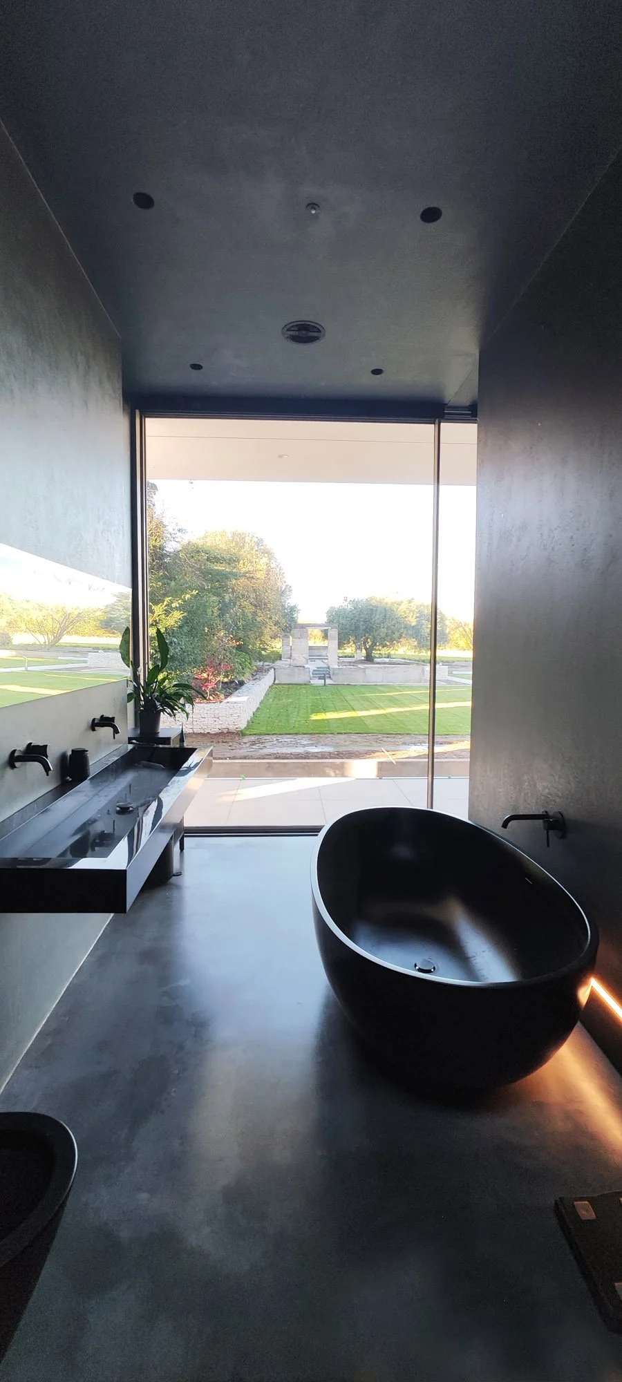 Modern bathroom with a dark freestanding bathtub, a wall-mounted sink with two faucets, a potted plant, and a large window view of a landscaped outdoor area with trees, grass, and stone features.