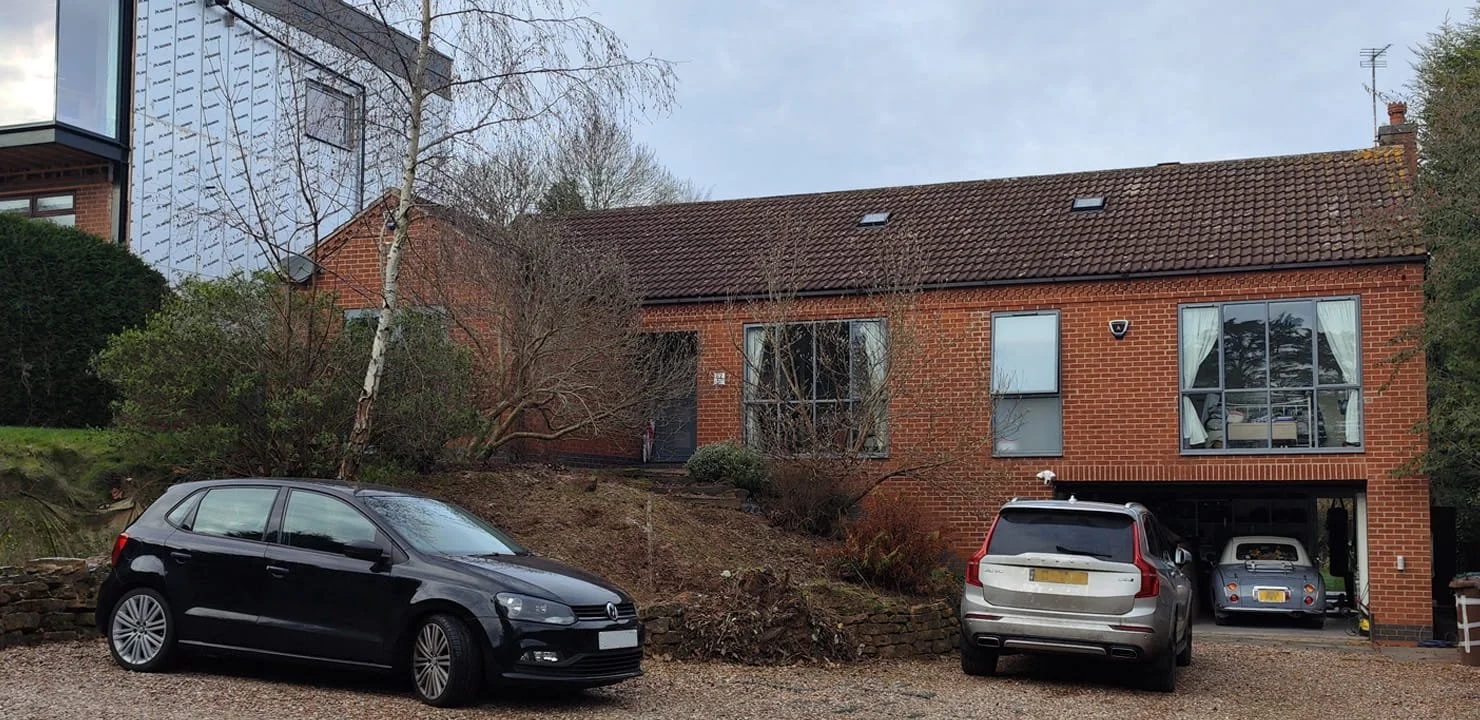 Residential house with brick exterior, garage, and driveway. Three cars are parked outside, and there are trees and shrubs around the house.