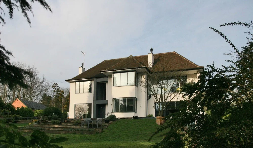A large two-story white house with a brown tiled roof, multiple large windows, and surrounded by trees and landscaped yard.