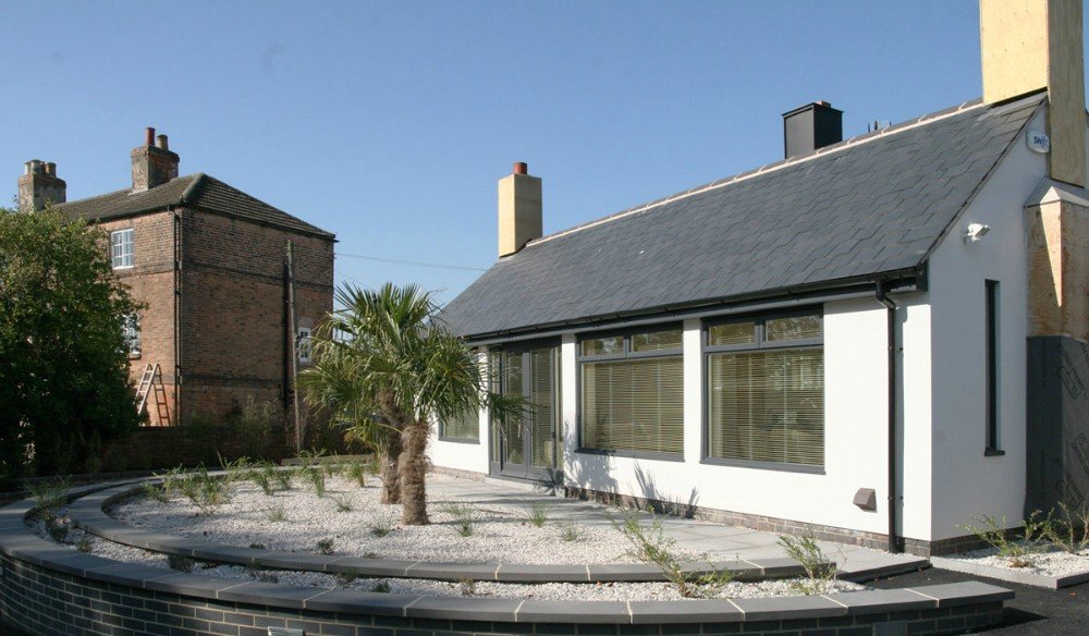 Modern white house with large windows, gray roof, and landscaped front yard with a small palm tree and ornamental plants.
