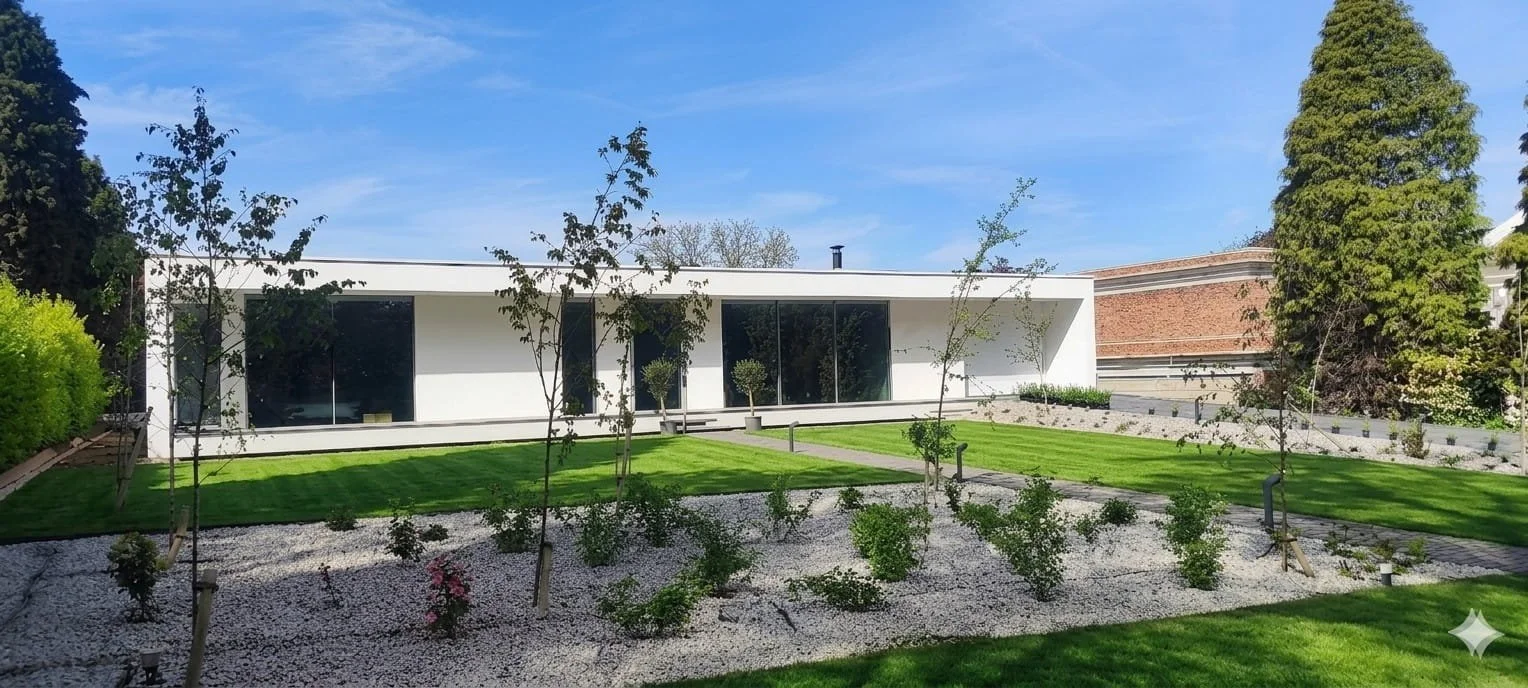 Modern white house with large glass windows, surrounded by a landscaped garden with neatly trimmed grass, young trees, and small bushes, under a blue sky.