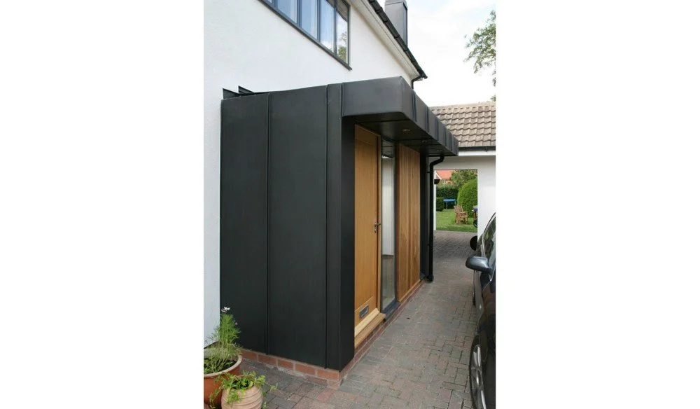 Exterior view of a house entrance with wooden door, black modern facade, and parked car nearby.