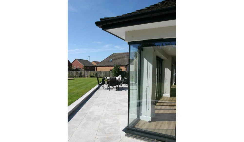 View of a backyard patio with a glass wall, outdoor dining table, and a lawn, with houses in the background under a clear blue sky.