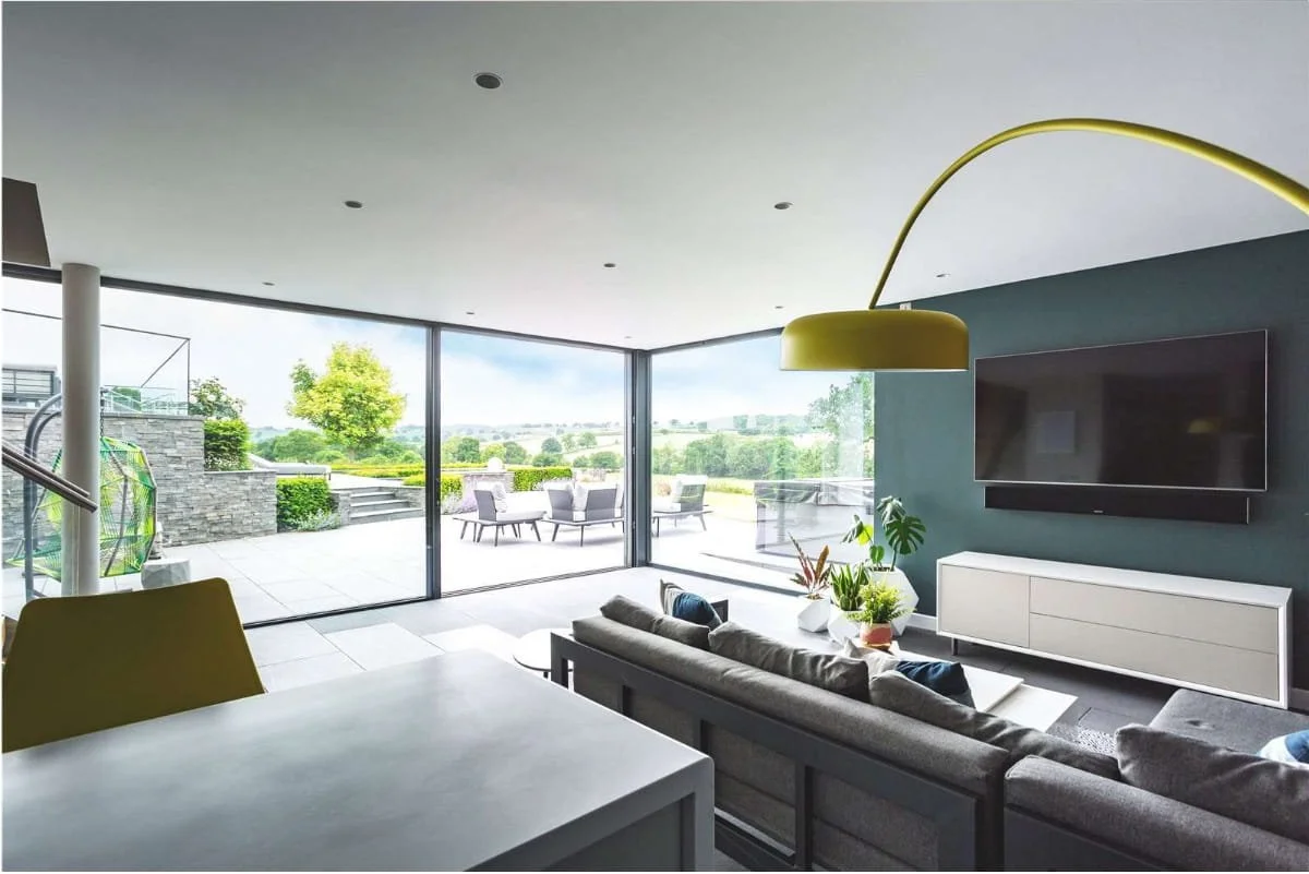 Modern living room with large glass sliding doors opening to an outdoor patio and scenic landscape, featuring a gray sofa, a coffee table with indoor plants, and a wall-mounted television.