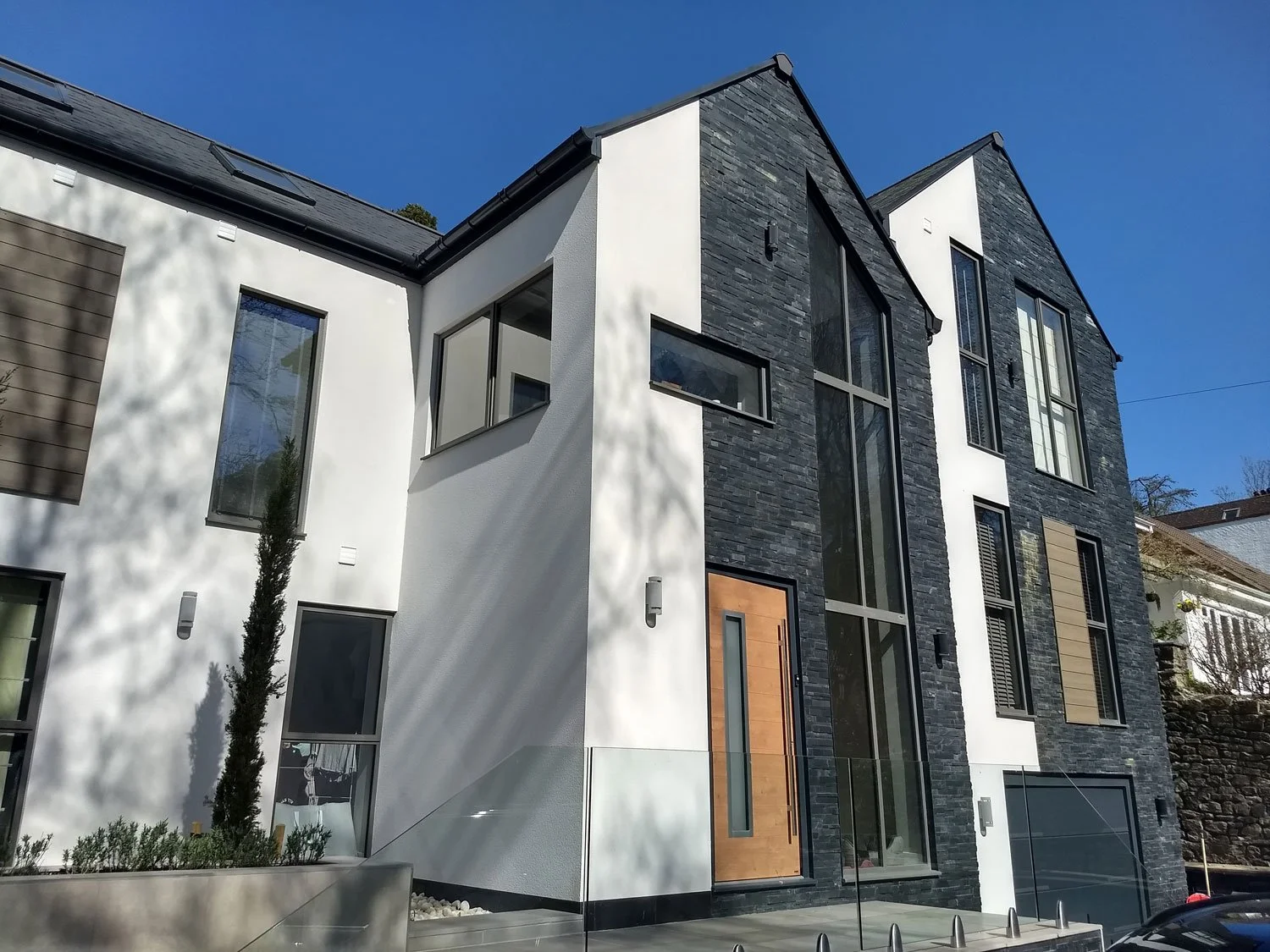 Modern multi-story house with black slate and white exterior walls, large windows, a wooden front door, and a glass railing, under a clear blue sky.