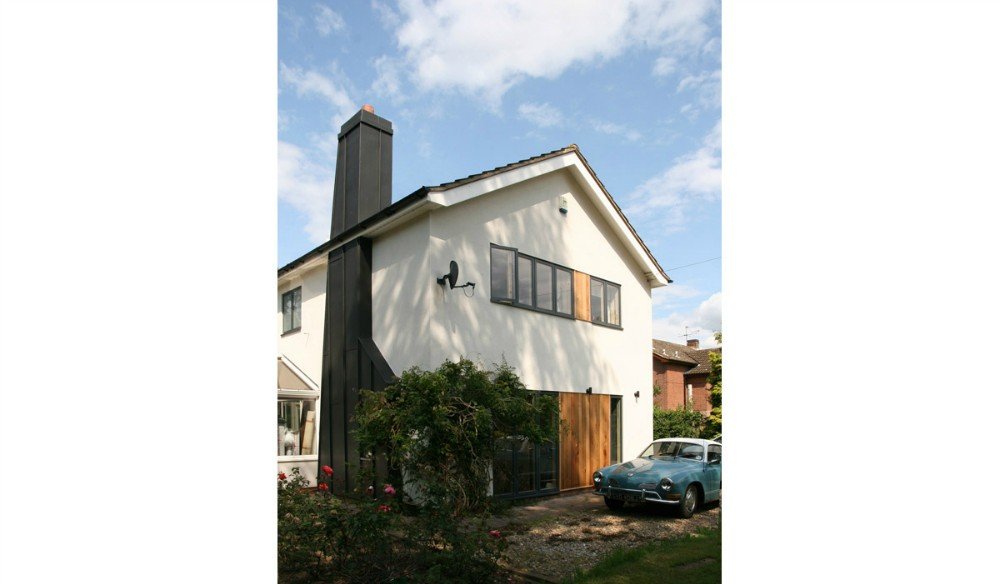 A two-story house with a white exterior, black chimney, and large front window, with a vintage car parked in front and a bush with pink flowers nearby.