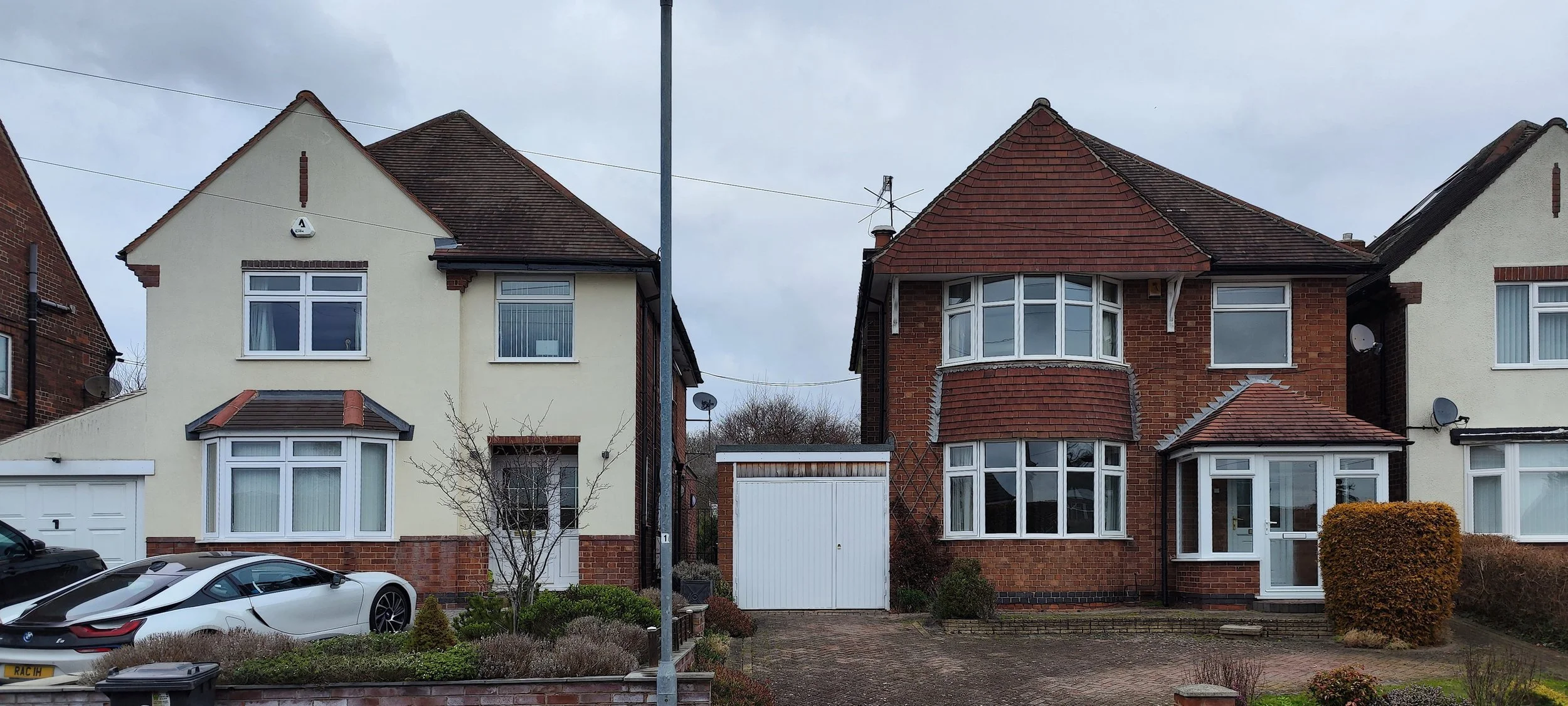 Two neighboring houses with parked cars in front, one house painted white and the other brick with a bay window, a driveway, and bushes.