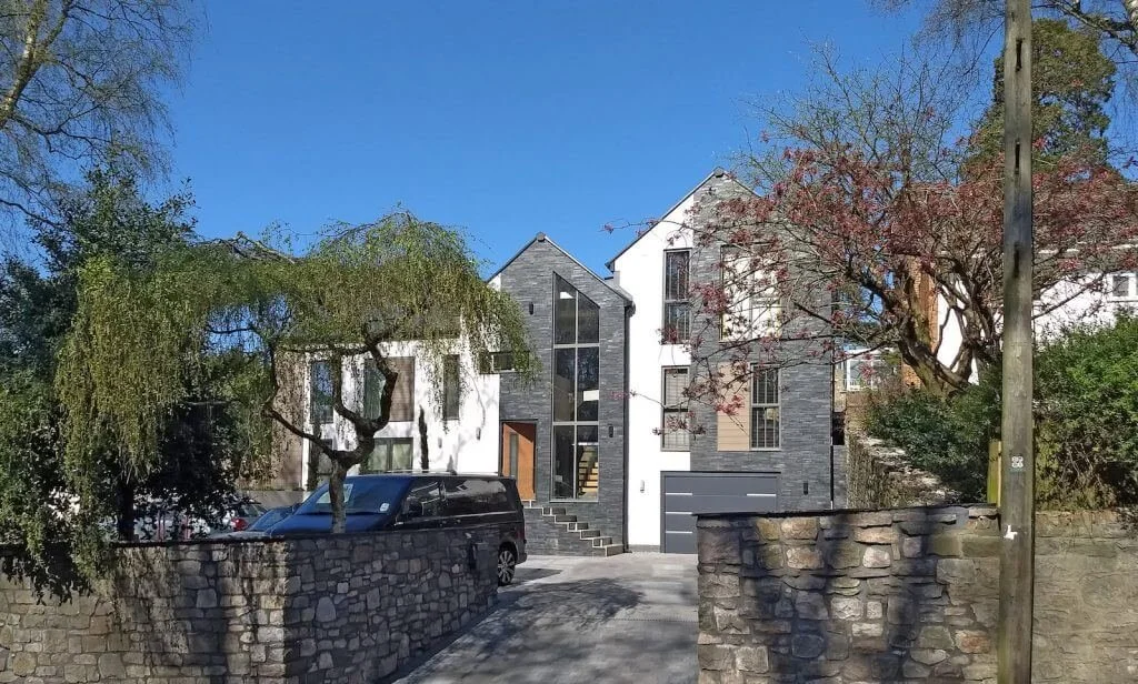Modern multi-story house with a mix of white and gray exterior, large windows, and a stone driveway, surrounded by trees and stone walls.