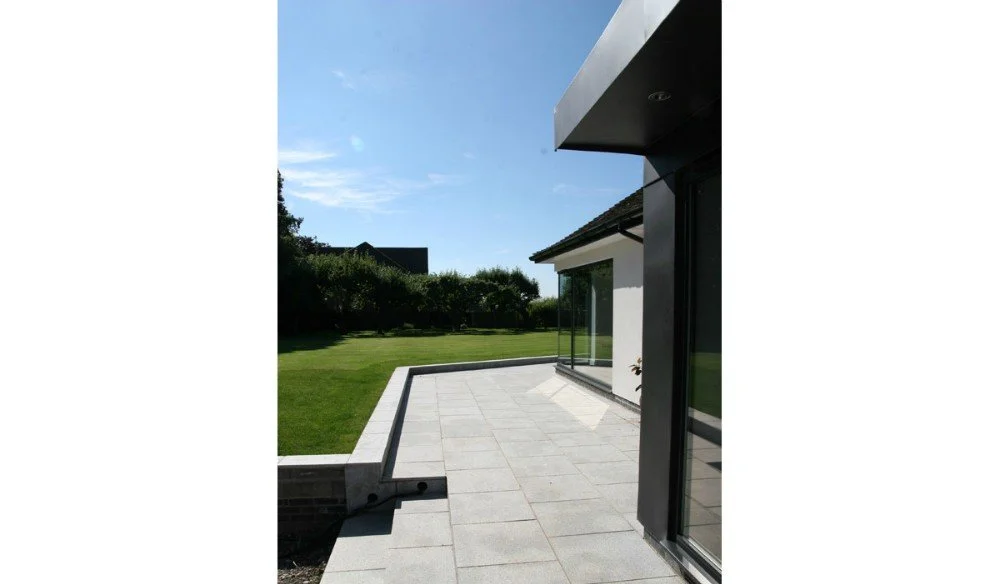 Modern house patio with large glass windows, concrete paving, and a grassy yard with trees in the background.