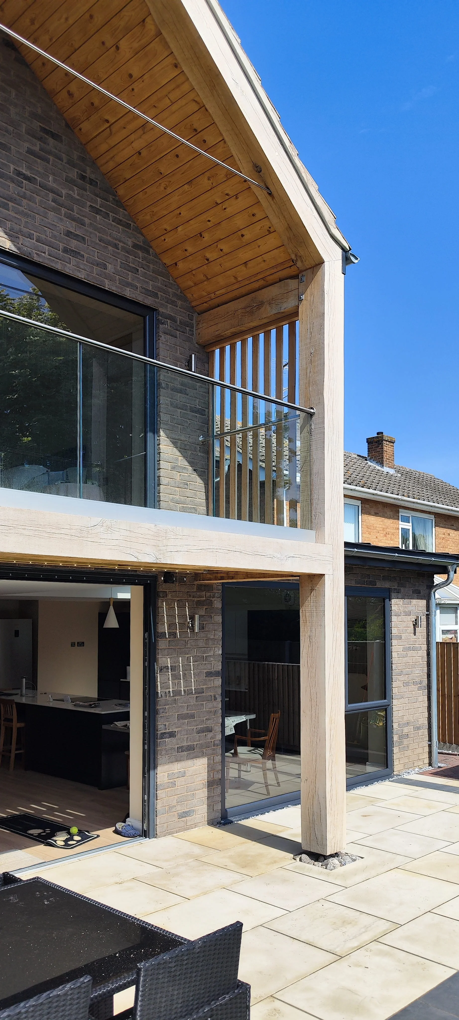 Exterior view of a modern two-story house with a brick facade, large sliding glass doors, and a balcony with glass railing. The patio features large beige tiles, a black outdoor table with chairs, and a swing set in the foreground. The sky is blue an