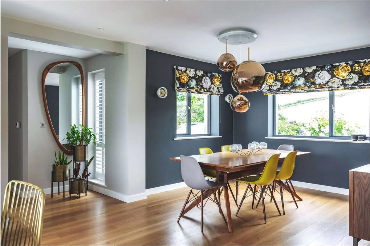 Dining room with a wooden table, six chairs in white and yellow, black walls, and large windows with floral valances. Modern copper pendant lights hang above the table. There are potted plants near a mirror on a white wall, and hardwood flooring.