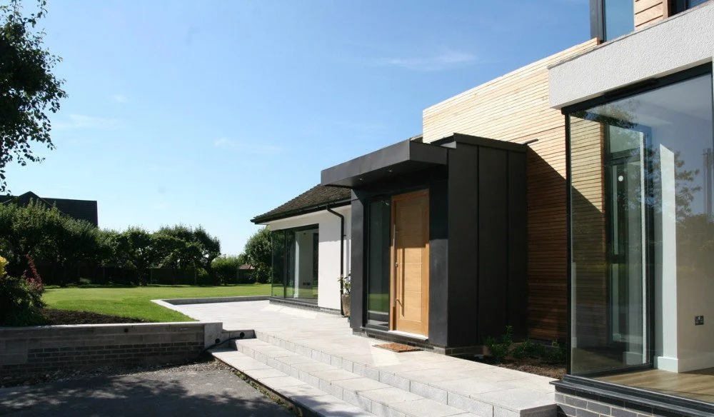 Modern house exterior with large glass windows, white walls, and wooden accents, surrounded by a well-maintained lawn and a paved walkway.