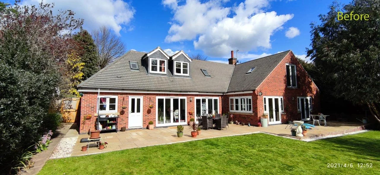 Backyard patio of a brick house before renovation. The patio has potted plants, outdoor furniture, and garden tools, with a grassy lawn in the foreground. The house has white-framed glass doors, windows, and roof dormers.