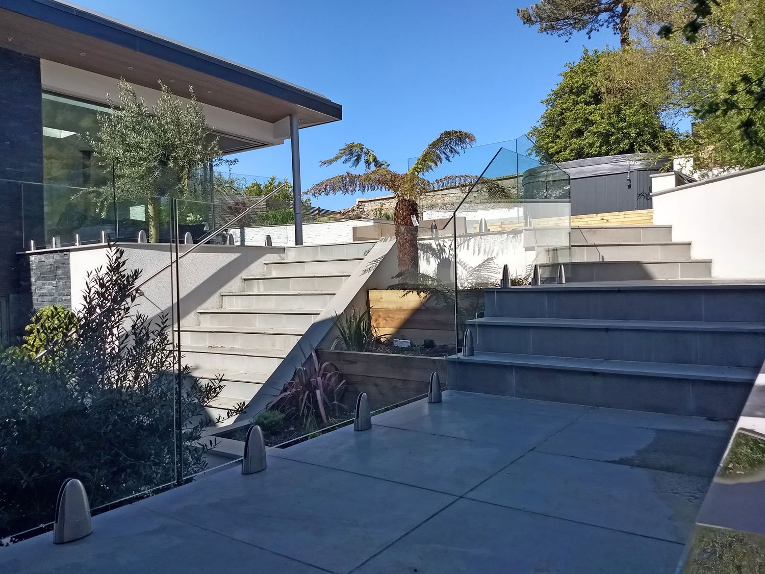 Modern outdoor staircase and patio area with glass railing, plants, and a clear blue sky.