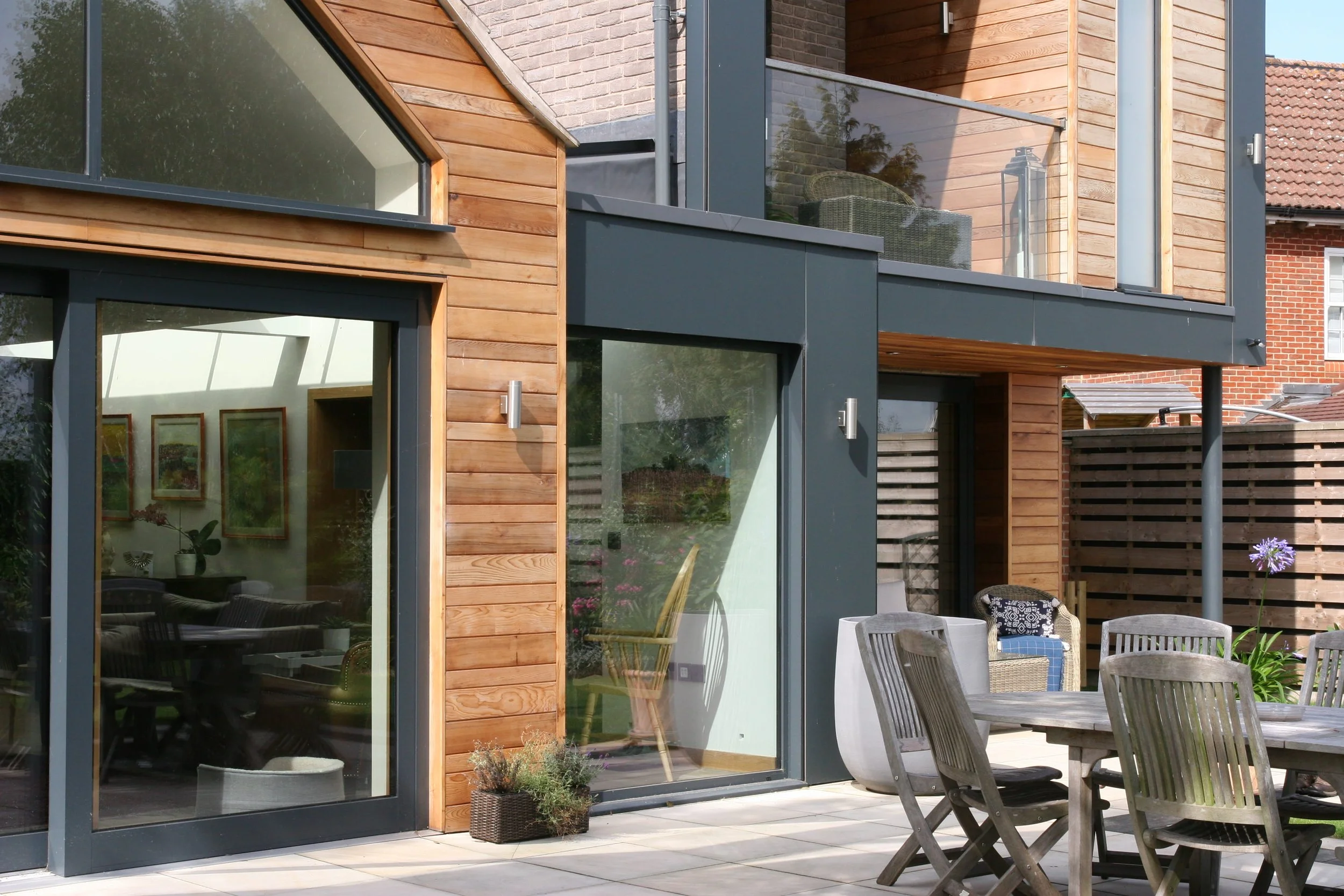 Modern house with large glass sliding doors, wood siding, and a balcony. There is an outdoor patio with dining furniture and a potted plant.
