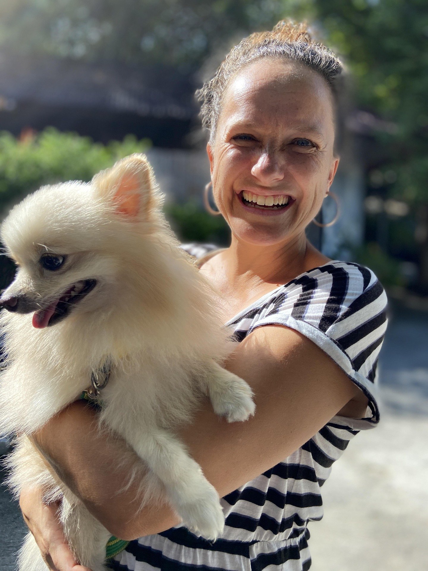 A woman smiling and holding a fluffy white dog outdoors with a blurred background of greenery and water.