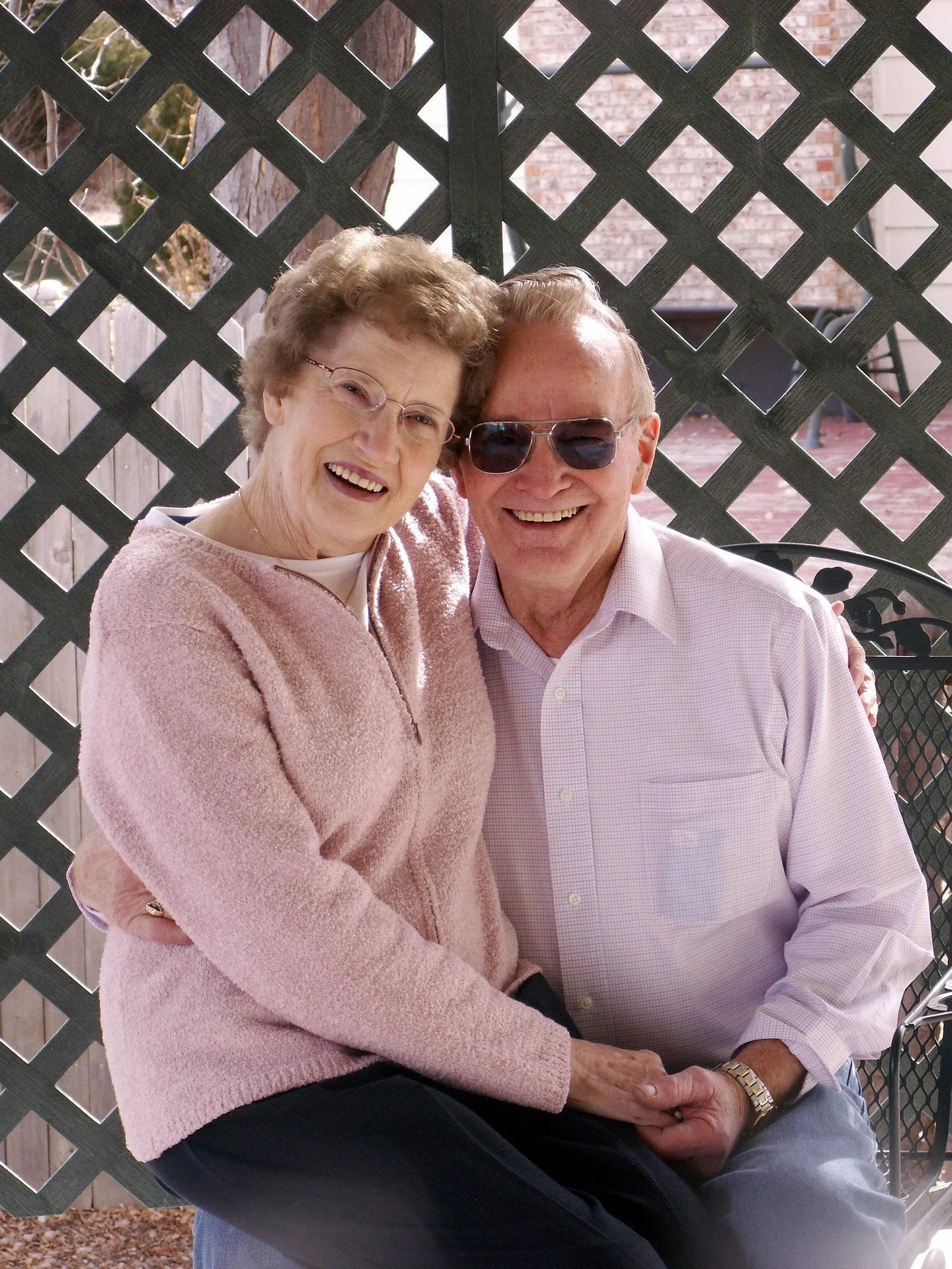 An elderly couple sitting close together on a bench outdoors, smiling happily, with a lattice fence and brick wall behind them.
