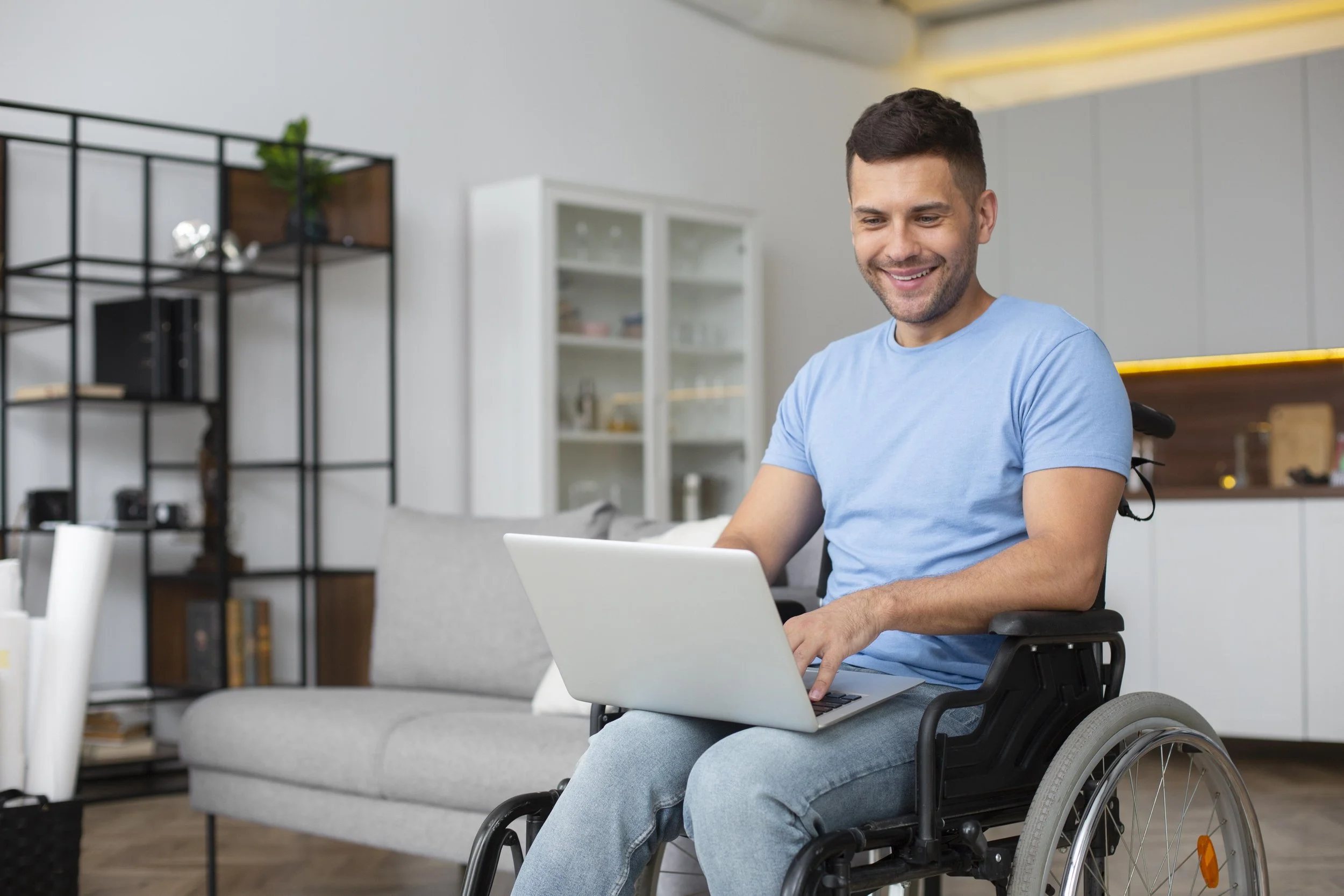 A smiling man in a wheelchair working on a laptop in a modern living room.