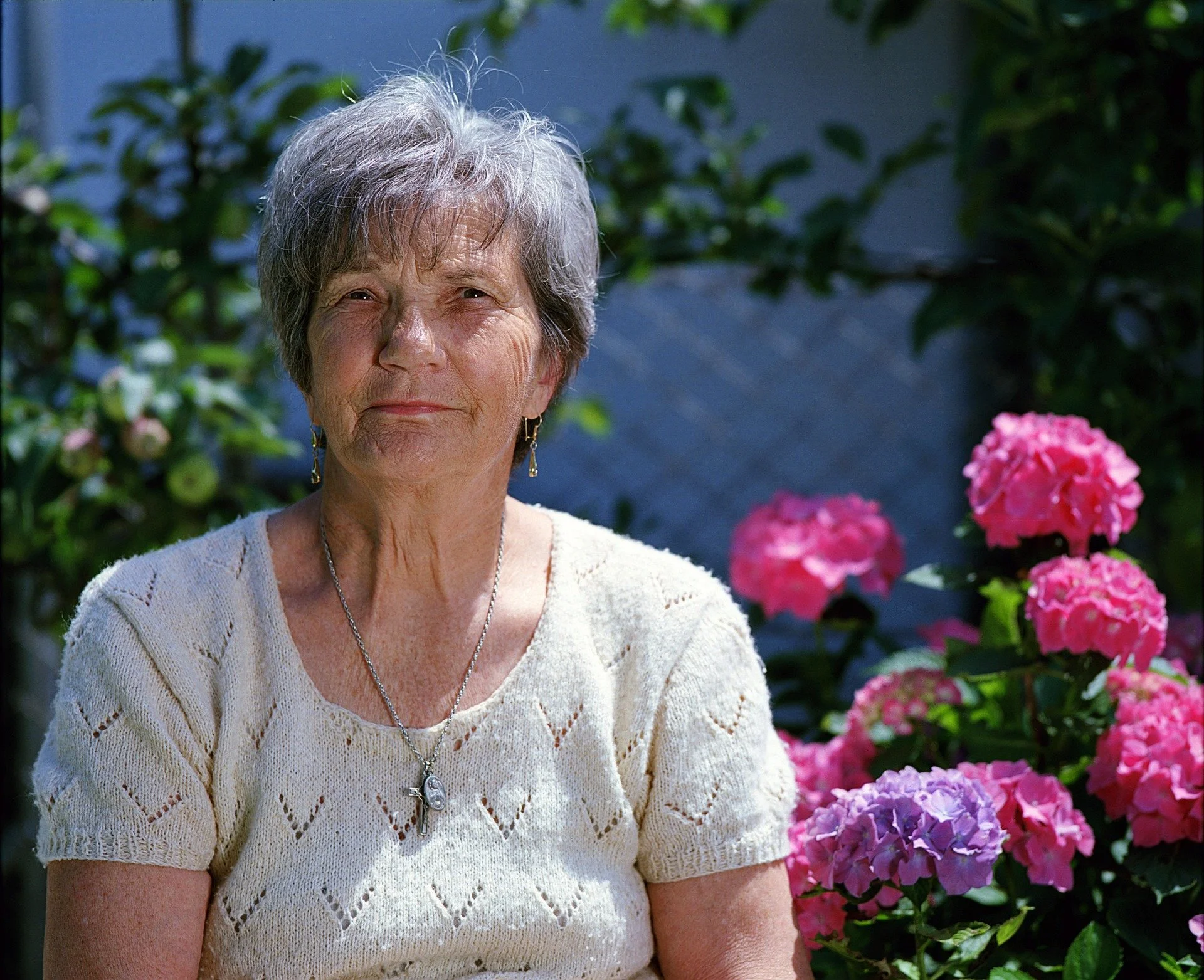An elderly woman with gray hair and earrings, wearing a cream-colored crocheted top with a necklace, standing outdoors with pink hydrangea flowers and green foliage in the background.