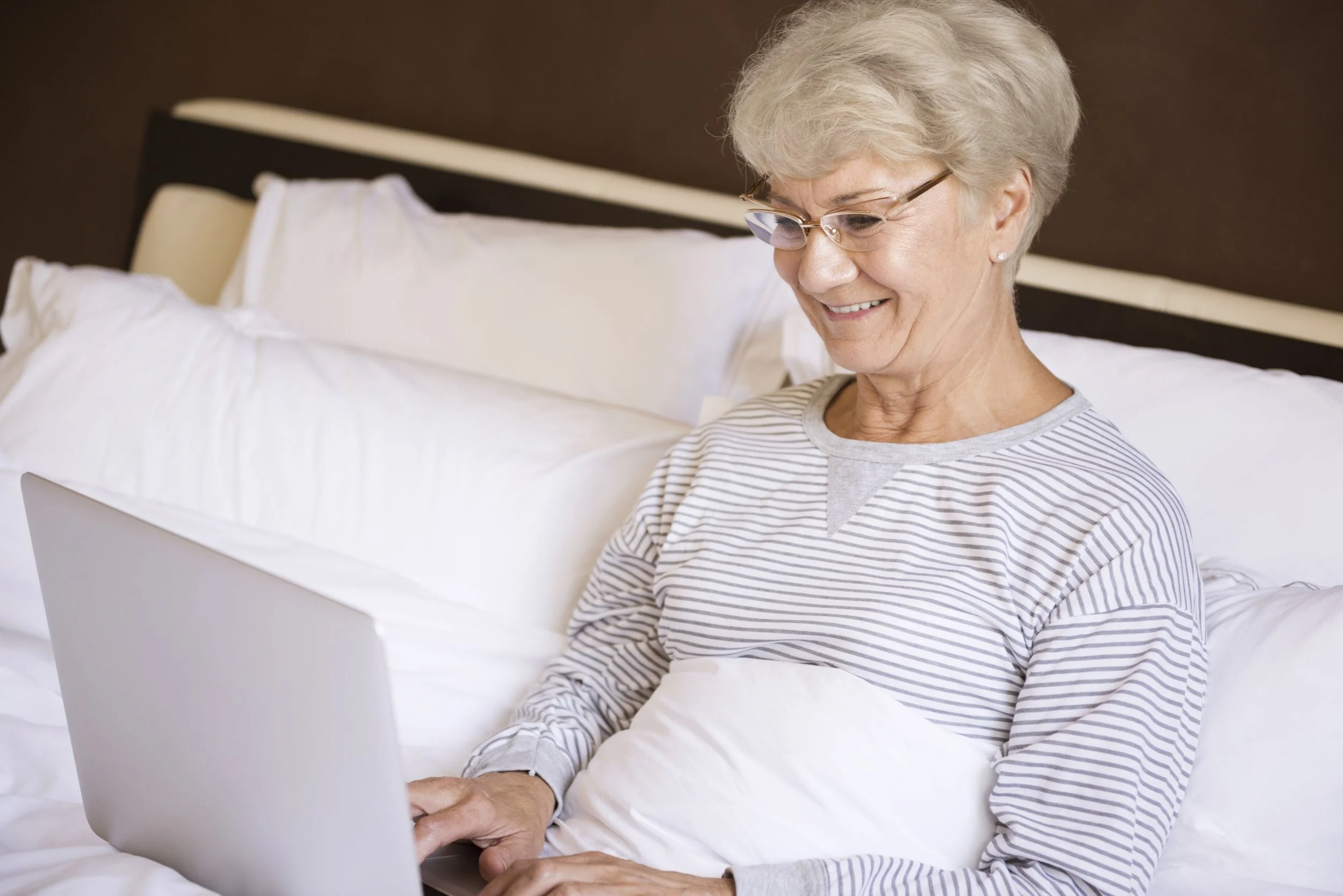 An elderly woman with gray hair, wearing glasses and a striped shirt, smiling while using a laptop in bed with white pillows and a dark headboard.
