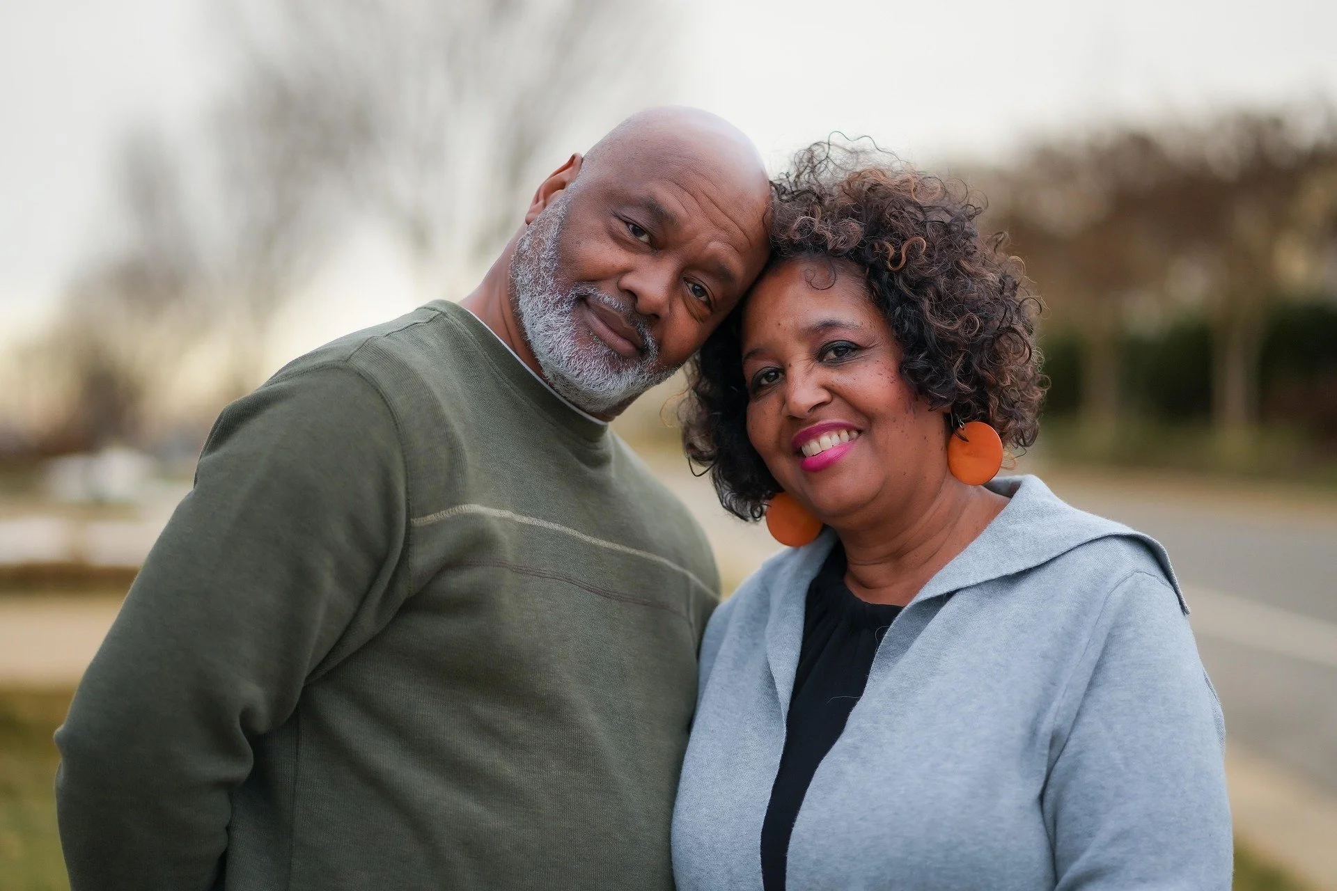 A smiling older man with a beard and bald head, leaning his head on an older woman with curly hair and bright orange earrings, outdoors with blurred trees and sky in the background.