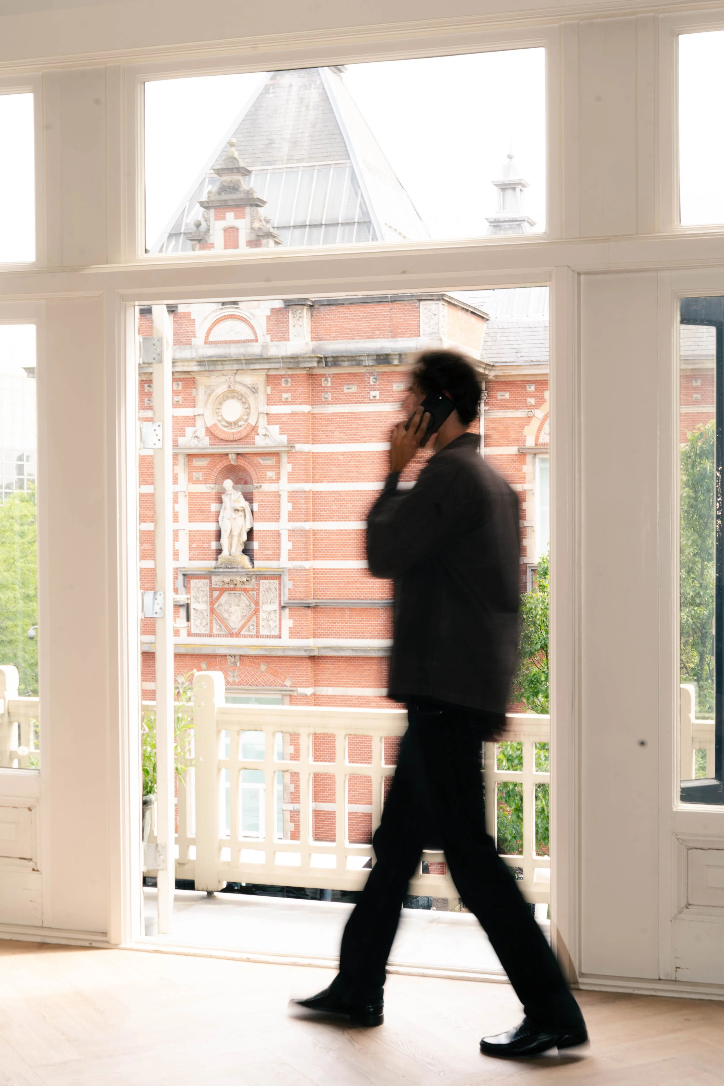 A man in a black suit walking inside a room with a large window showing a brick building and a balcony outside.