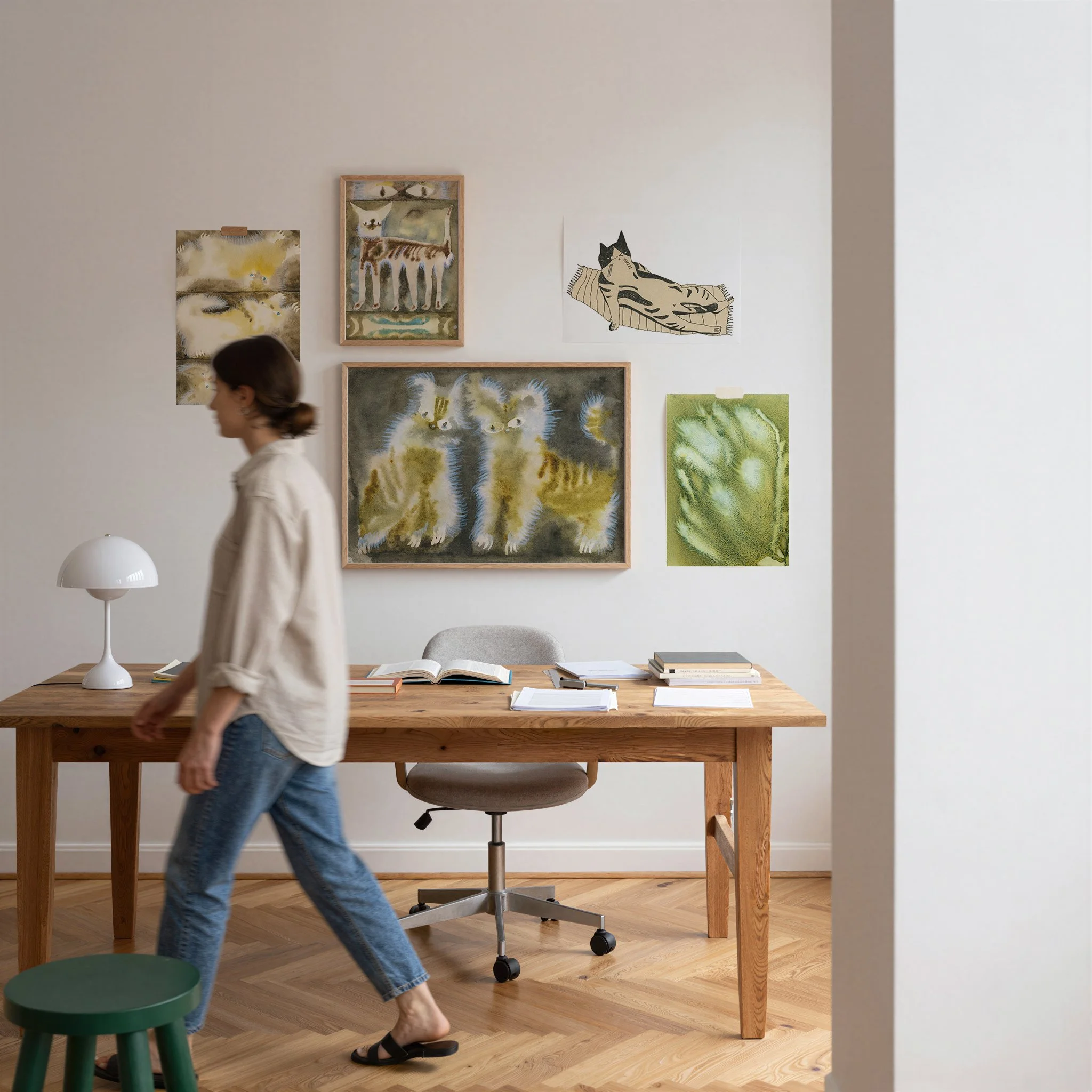 A woman walking through a room with artwork on the wall, a wooden desk with books and papers, a white modern table lamp, and a green stool. The room has wooden flooring and white walls.