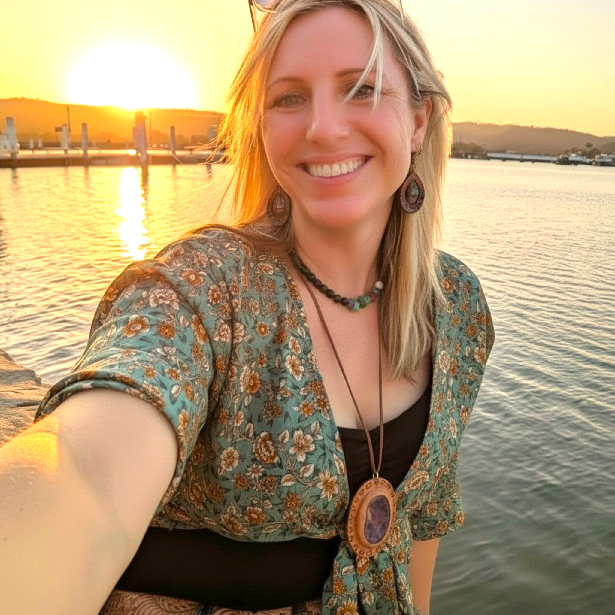 A woman smiling at sunset near a body of water, wearing a floral top, pendant necklace, and earrings.