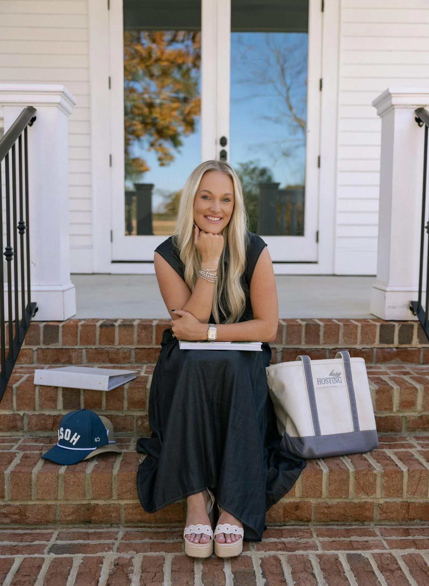 A woman sitting on brick steps outside a house, smiling at the camera, holding a closed laptop, with a tote bag and baseball cap nearby.