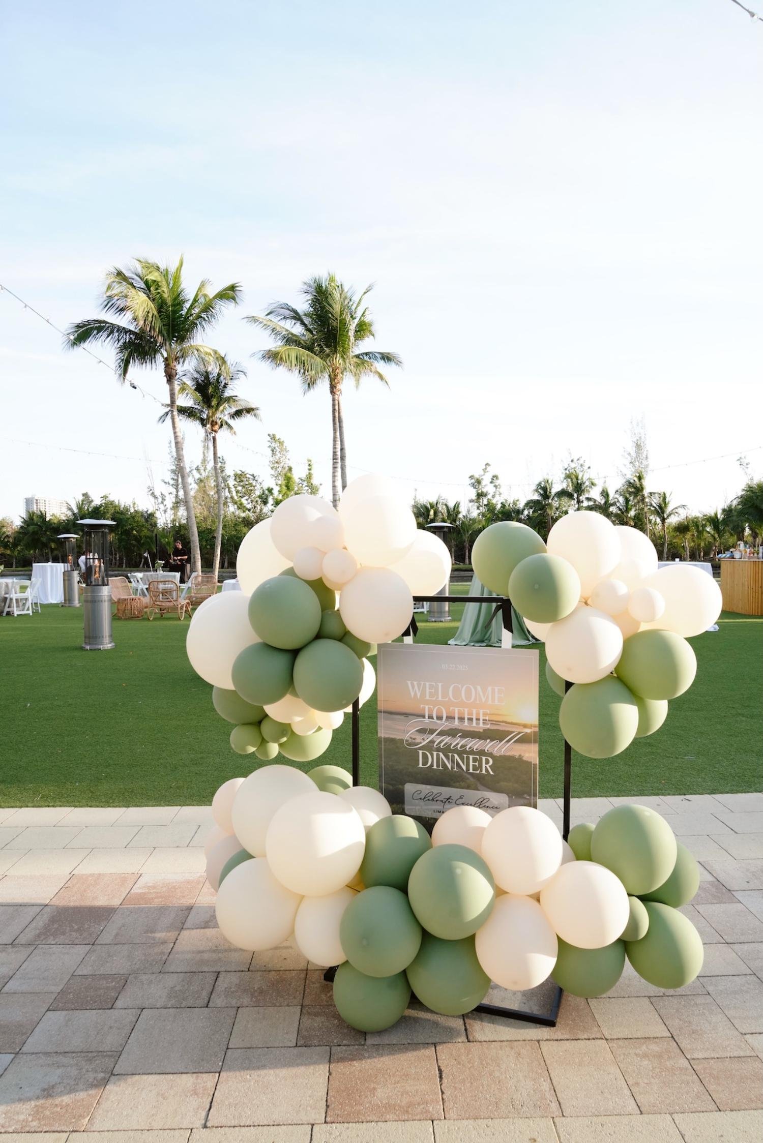 Decorative balloon arch with a sign welcoming guests to a farewell dinner, set outdoors with palm trees and tables in the background.
