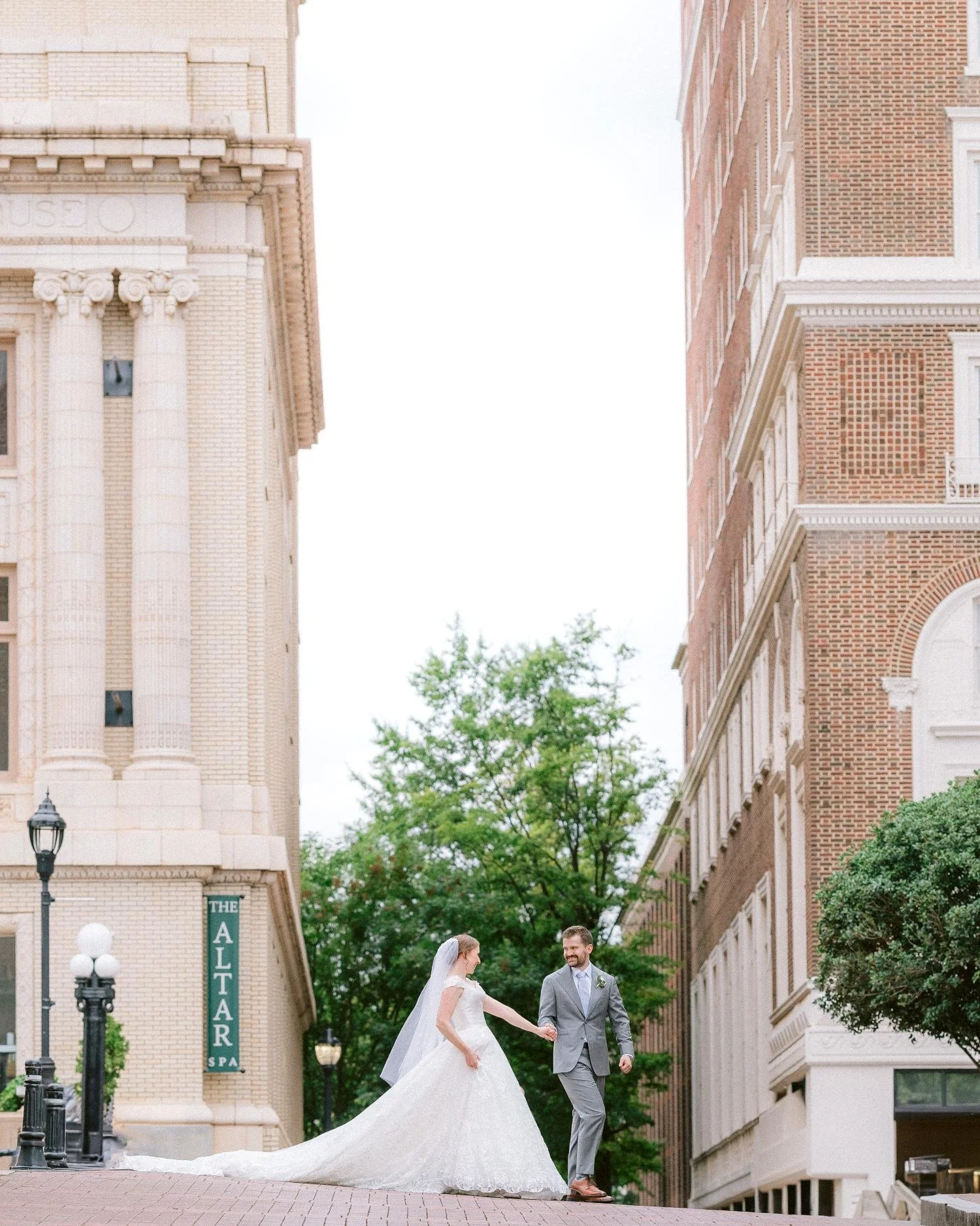 Bride and groom holding hands and walking on a city street between tall brick and stone buildings with trees in the background.