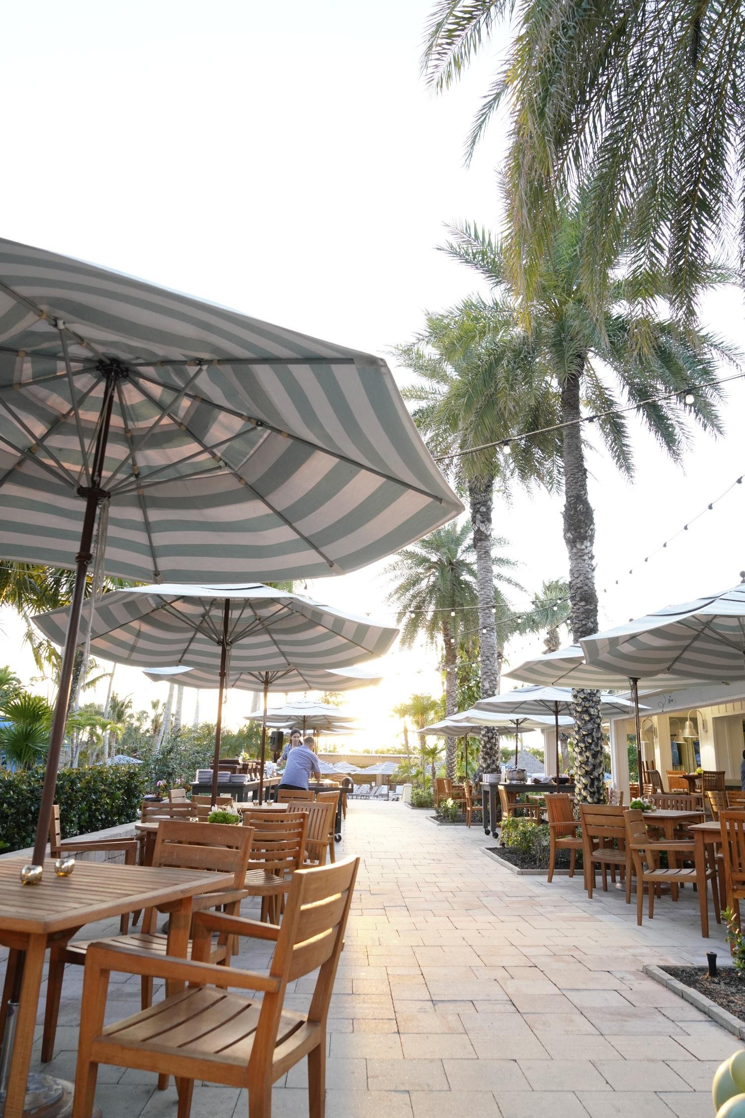Outdoor patio with wooden tables and chairs, striped umbrellas, tall palm trees, and a person setting up a buffet at a restaurant in the evening.