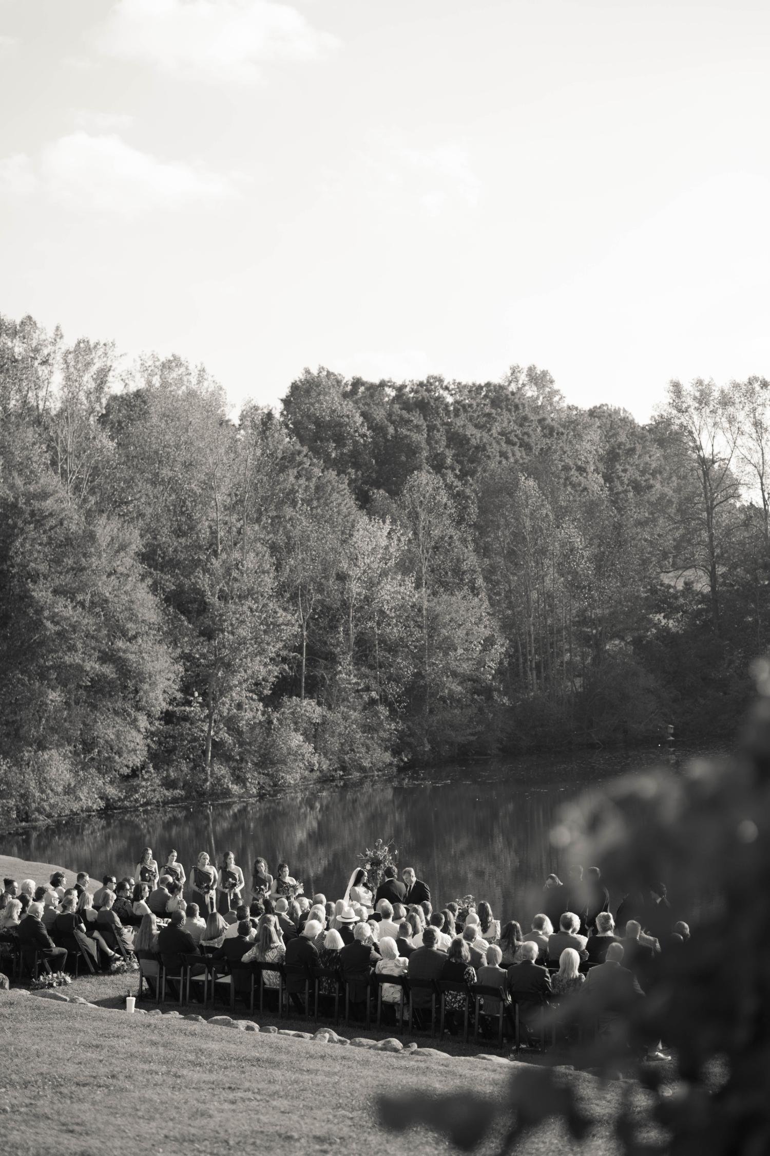 A black-and-white photo of an outdoor wedding ceremony by a lakeside, with guests seated and a wedding party standing in front of a scenic wooded backdrop.