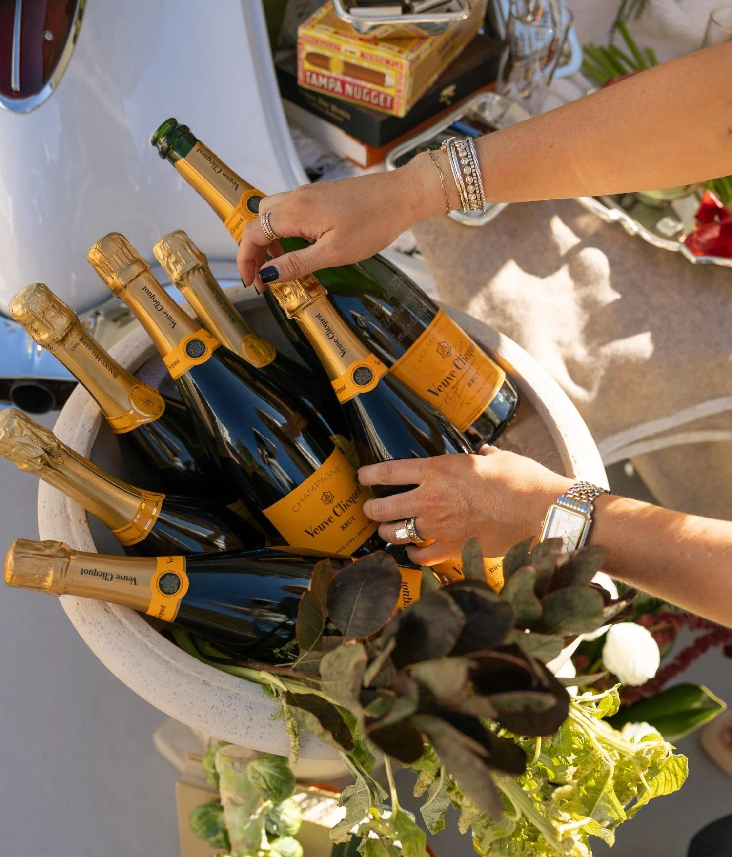 Hands reaching for bottles of Veuve Clicquot champagne in a large white cooler at an outdoor event.