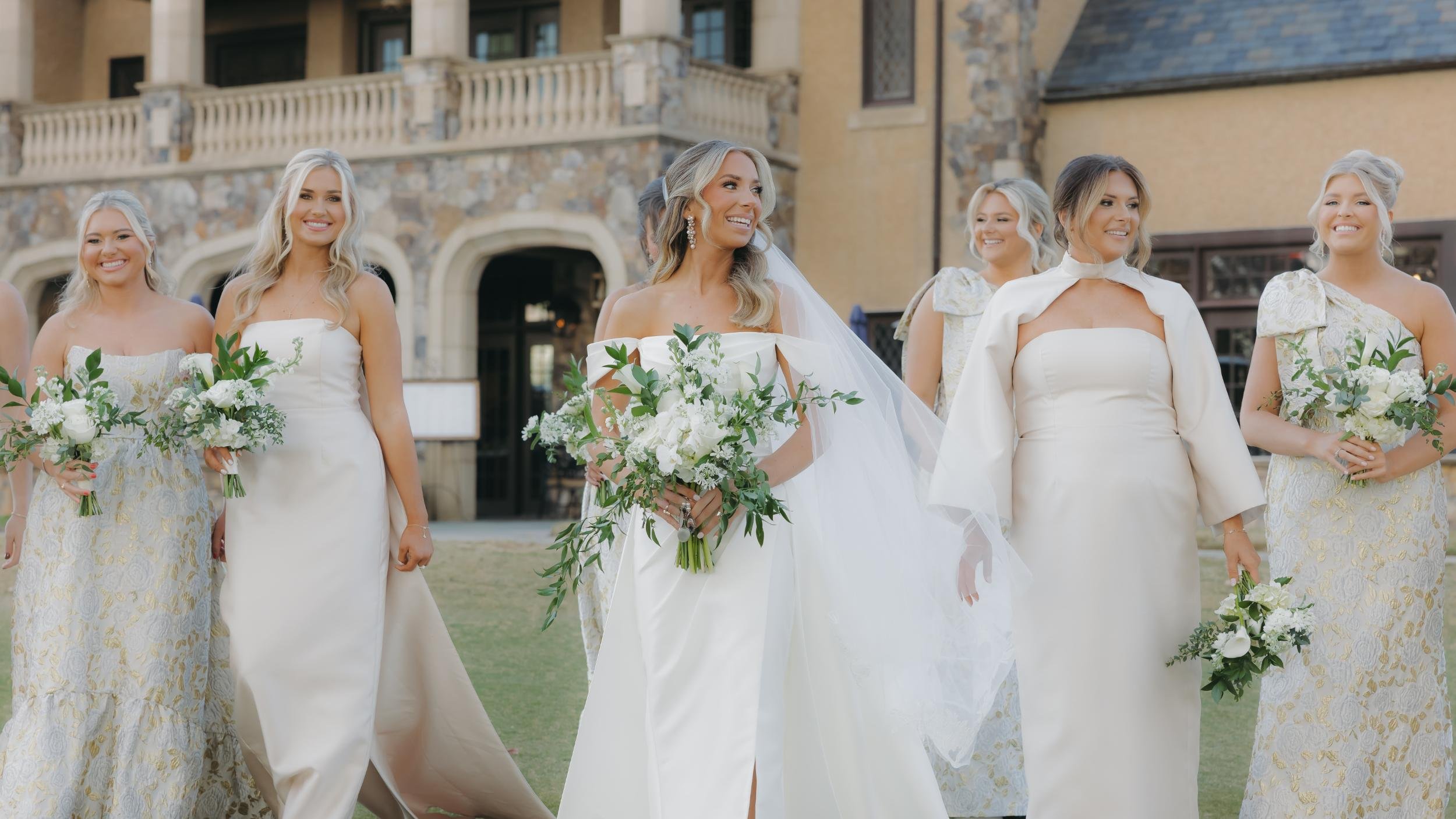 A bride and her bridesmaids walk outside on a grassy area in front of a large stone and stucco building. The bride is wearing an off-the-shoulder white wedding dress with a veil and holding a bouquet of white flowers and greenery. The bridesmaids are dressed in cream and gold gowns, holding matching bouquets of white flowers and greenery, smiling and enjoying the occasion.
