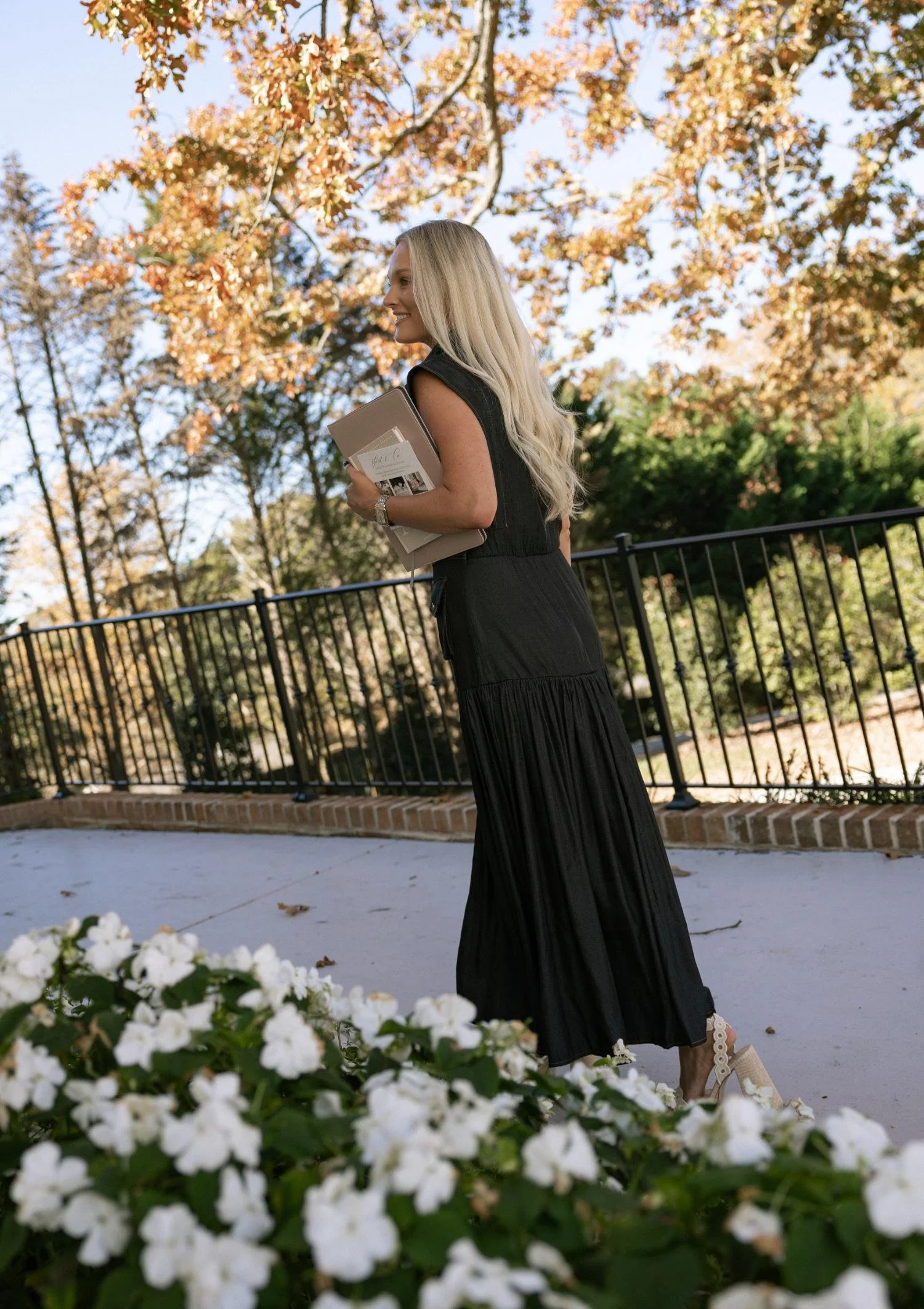 A woman with long blonde hair wearing a black dress walking outdoors on a pathway, holding a beige folder and a notebook, with trees and blooming white flowers in the background.