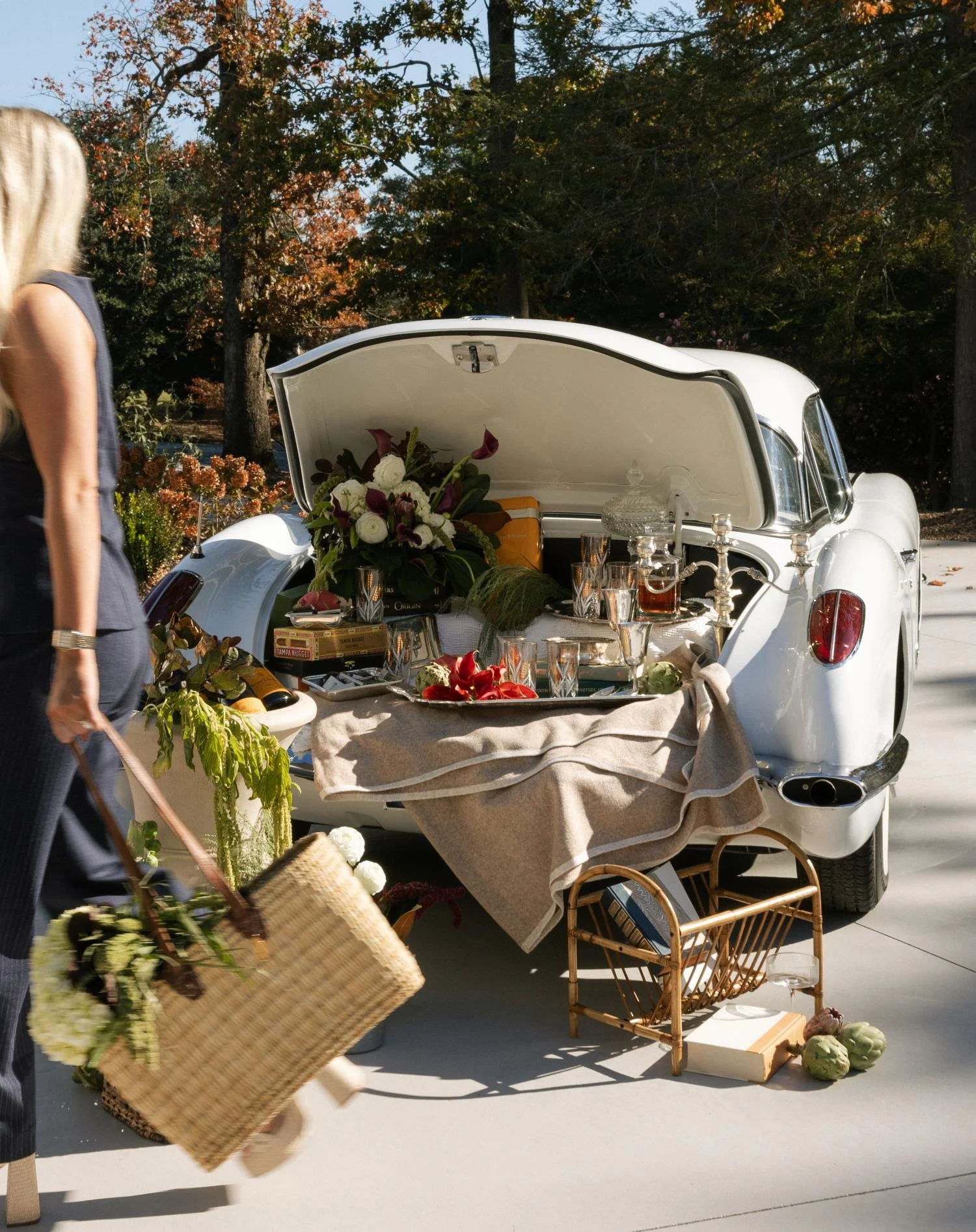 Vintage white car with its trunk open, filled with various items for a yard sale, including glassware, flowers, and boxes, set outside on a sunny day with trees in the background.
