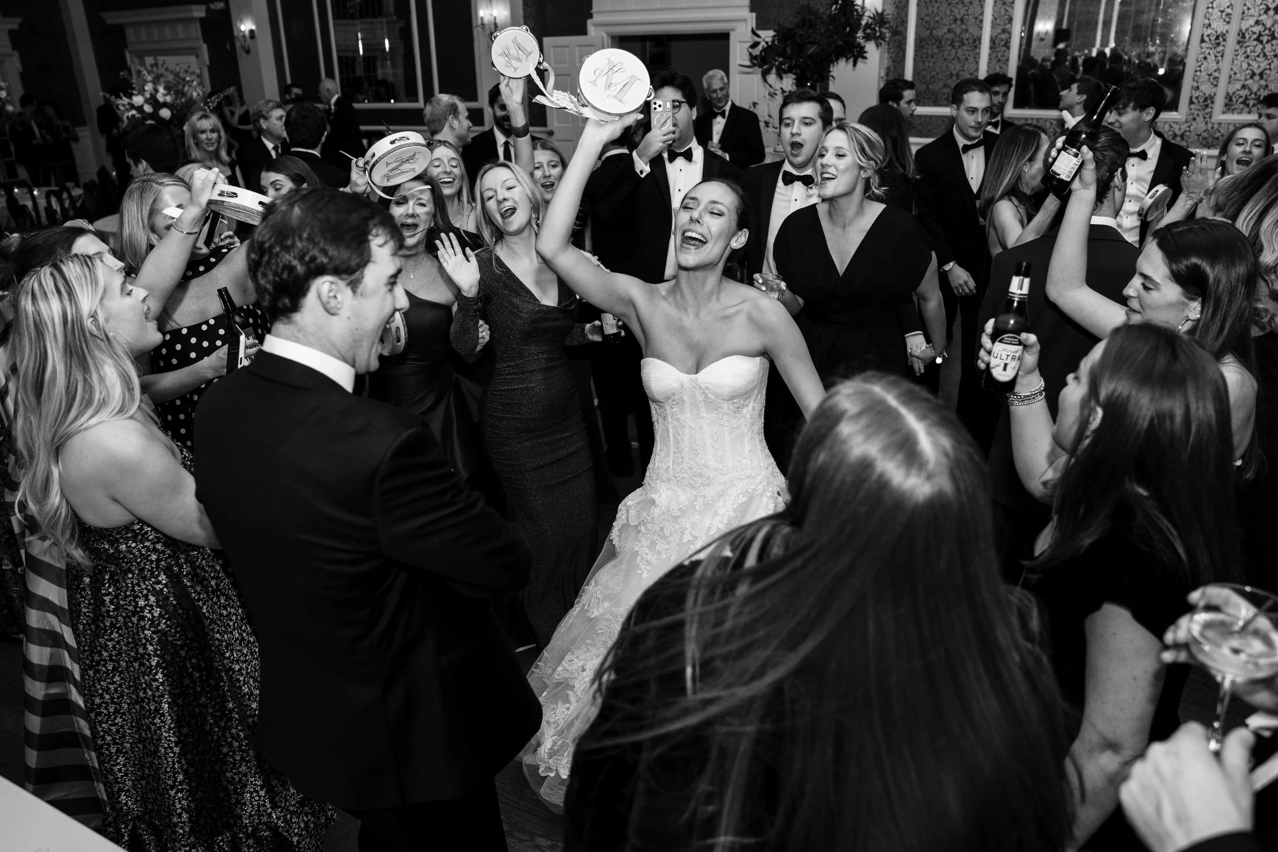 A black-and-white photo of a wedding reception with a bride dancing and celebrating with guests holding drinks and party favors.