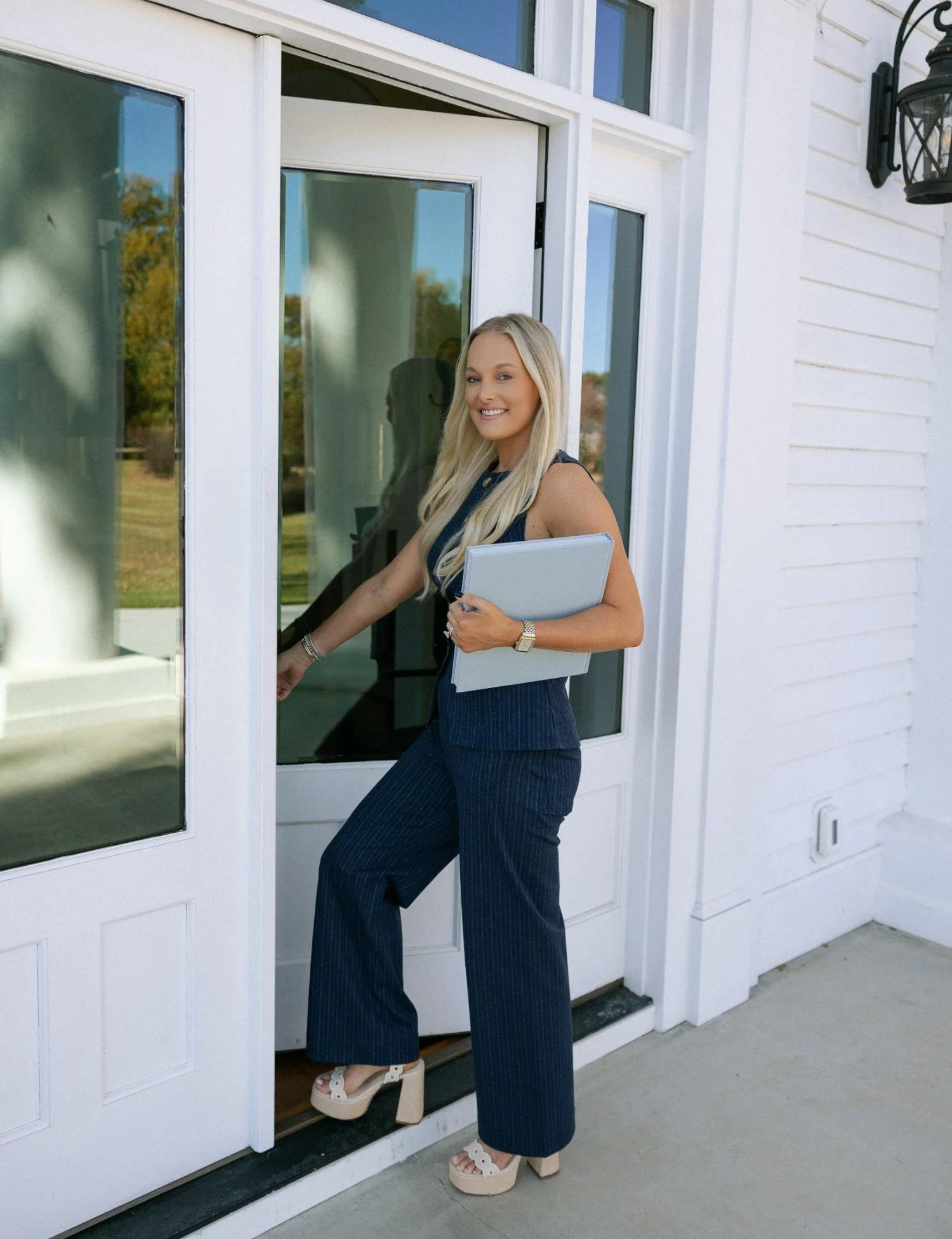 A professional woman with long blonde hair wearing a navy pinstripe suit and beige platform shoes, holding a folder, as she enters a house through a glass door.