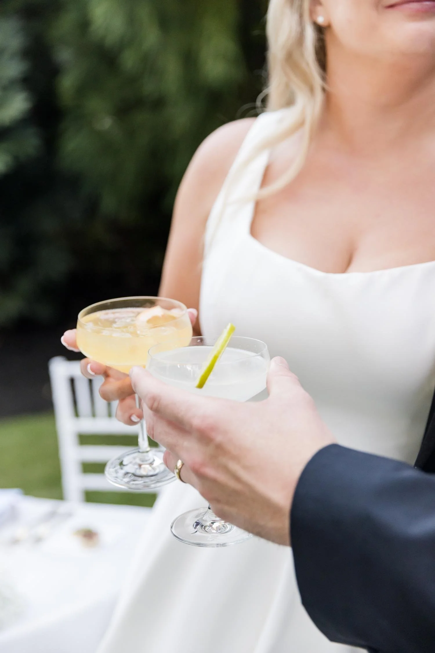 Close-up of a woman in a white dress holding two cocktails, one with a lime wedge, at an outdoor celebration.