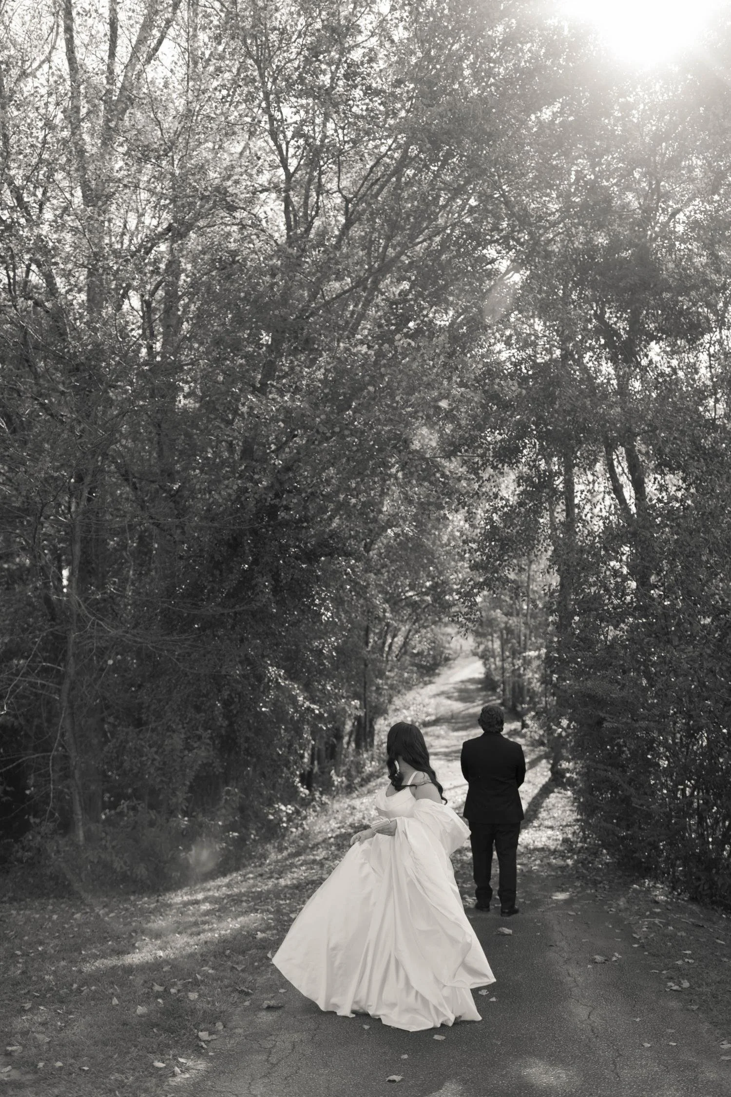 A black and white photo of a woman in a wedding dress and a man in a suit walking away on a forest path.