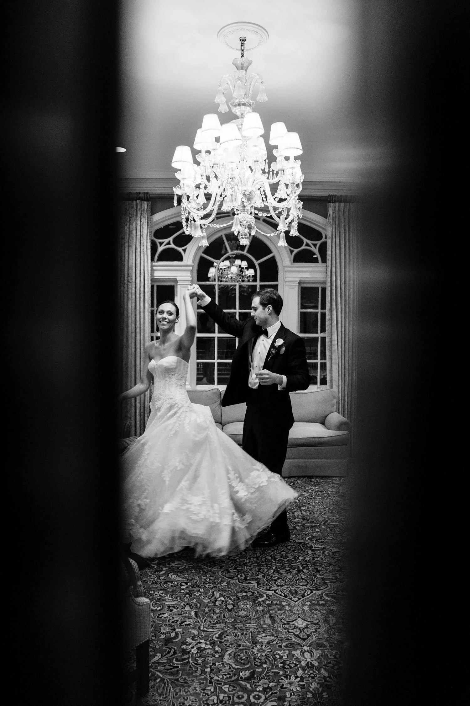 A bride and groom dancing in an elegant room with large windows, a chandelier, and ornate curtains, seen through a partially open door.