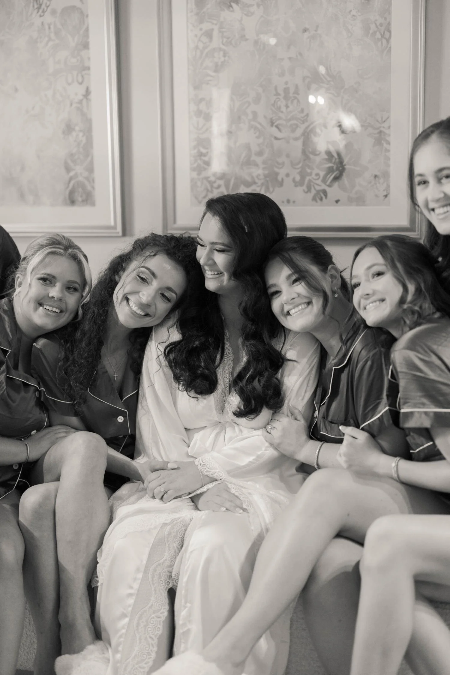 A group of women smiling and sitting close together, with one woman in a white robe at the center, likely getting ready for a special occasion.