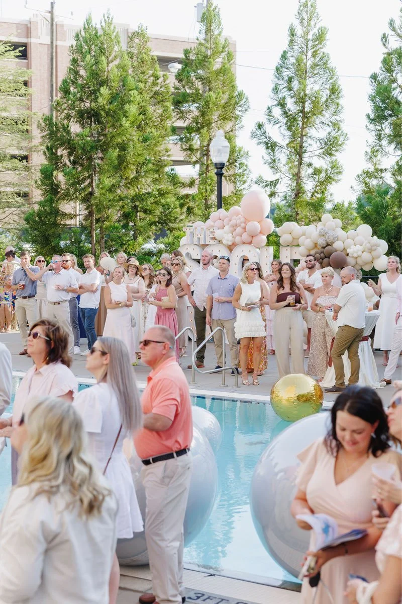 Group of people gathered around a pool at an outdoor event with balloon decorations and trees in the background.