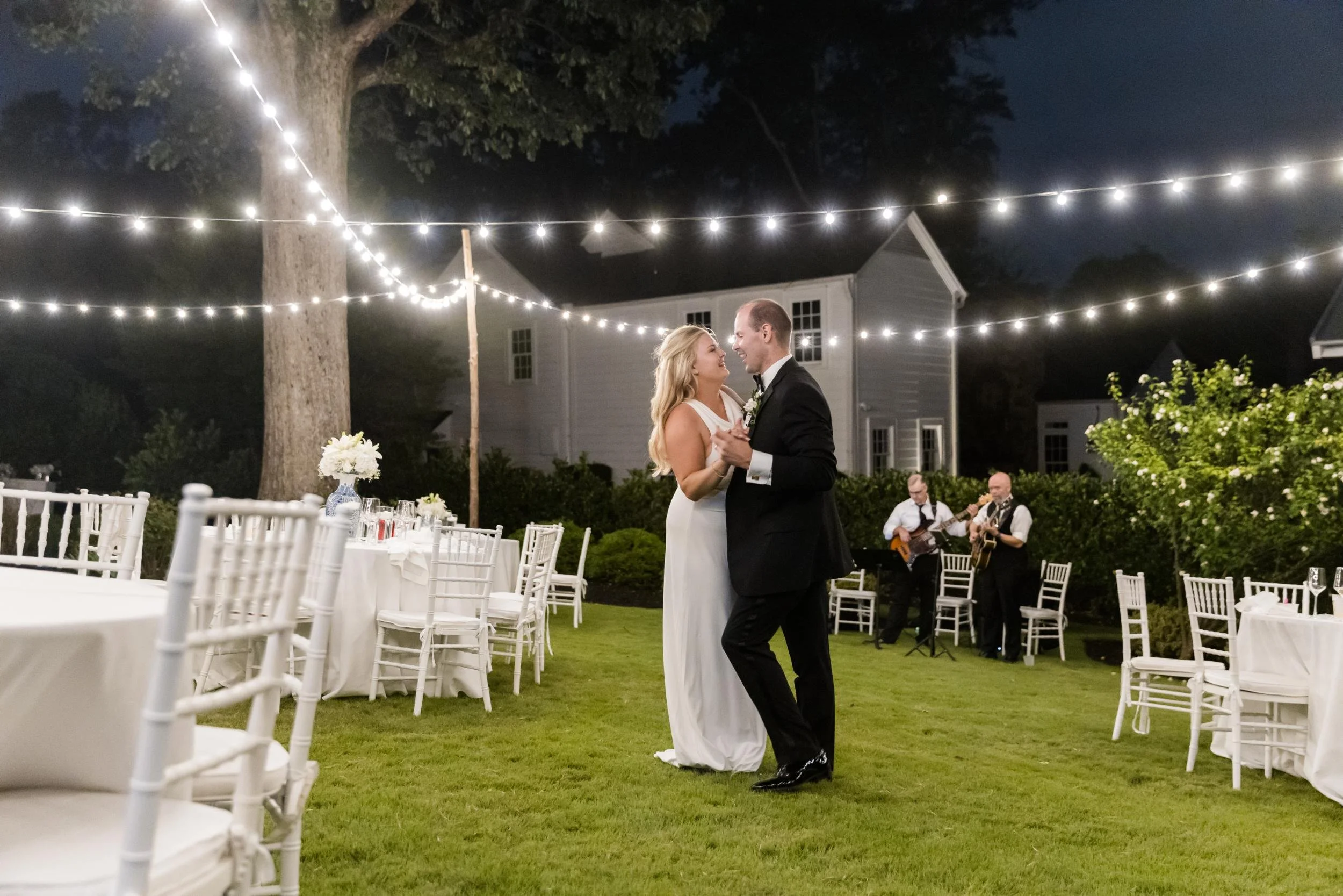 A bride and groom dancing outdoors at night under string lights on a decorated wedding reception setup with tables, chairs, and live musicians playing guitar in the background.